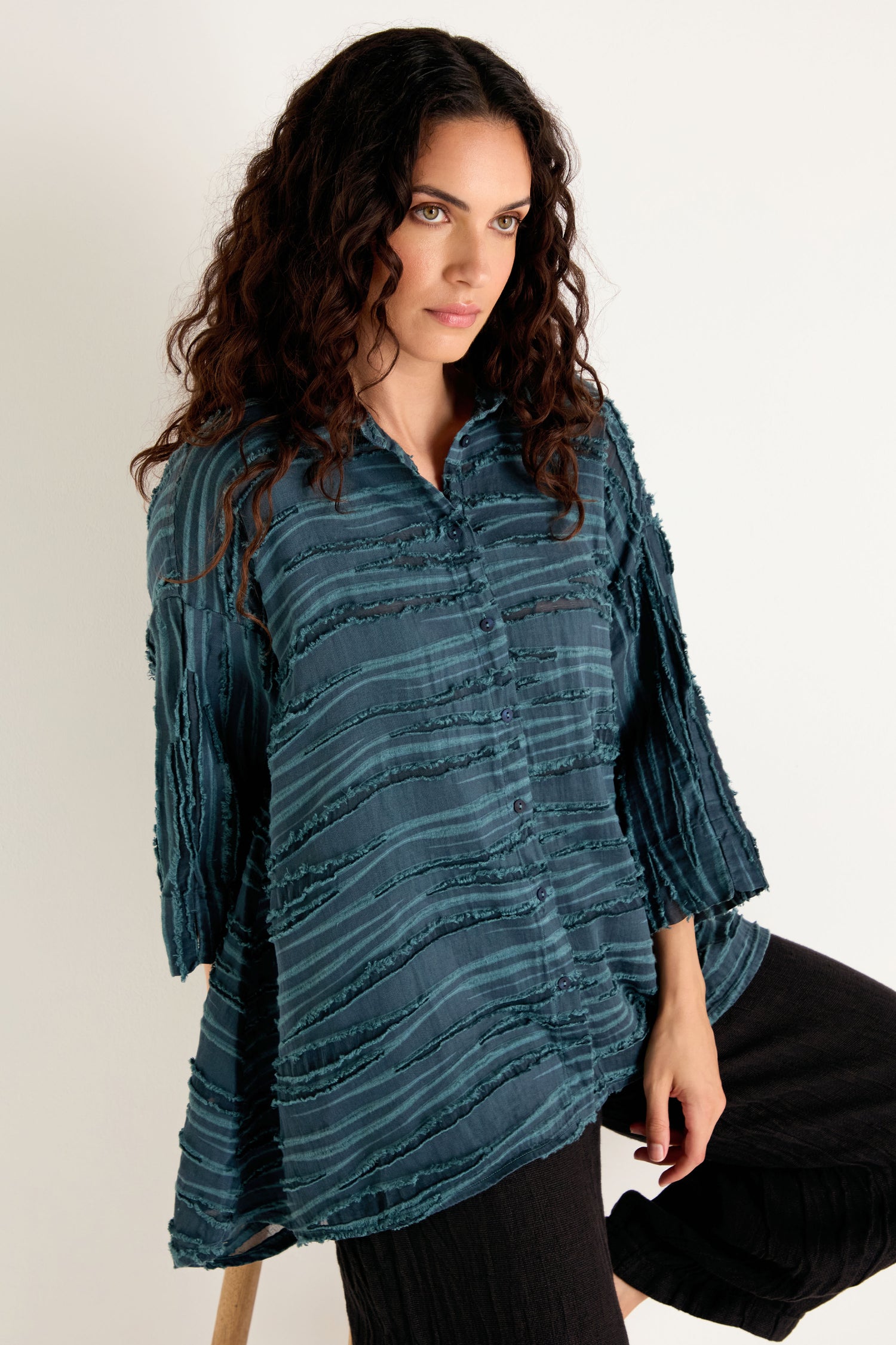 Woman with long curly hair wearing the Deep Turquoise Textured Stripe Flared Shirt—a linen silk cotton blend in a textured, button-up tunic style—paired with black pants, standing against a plain light background.