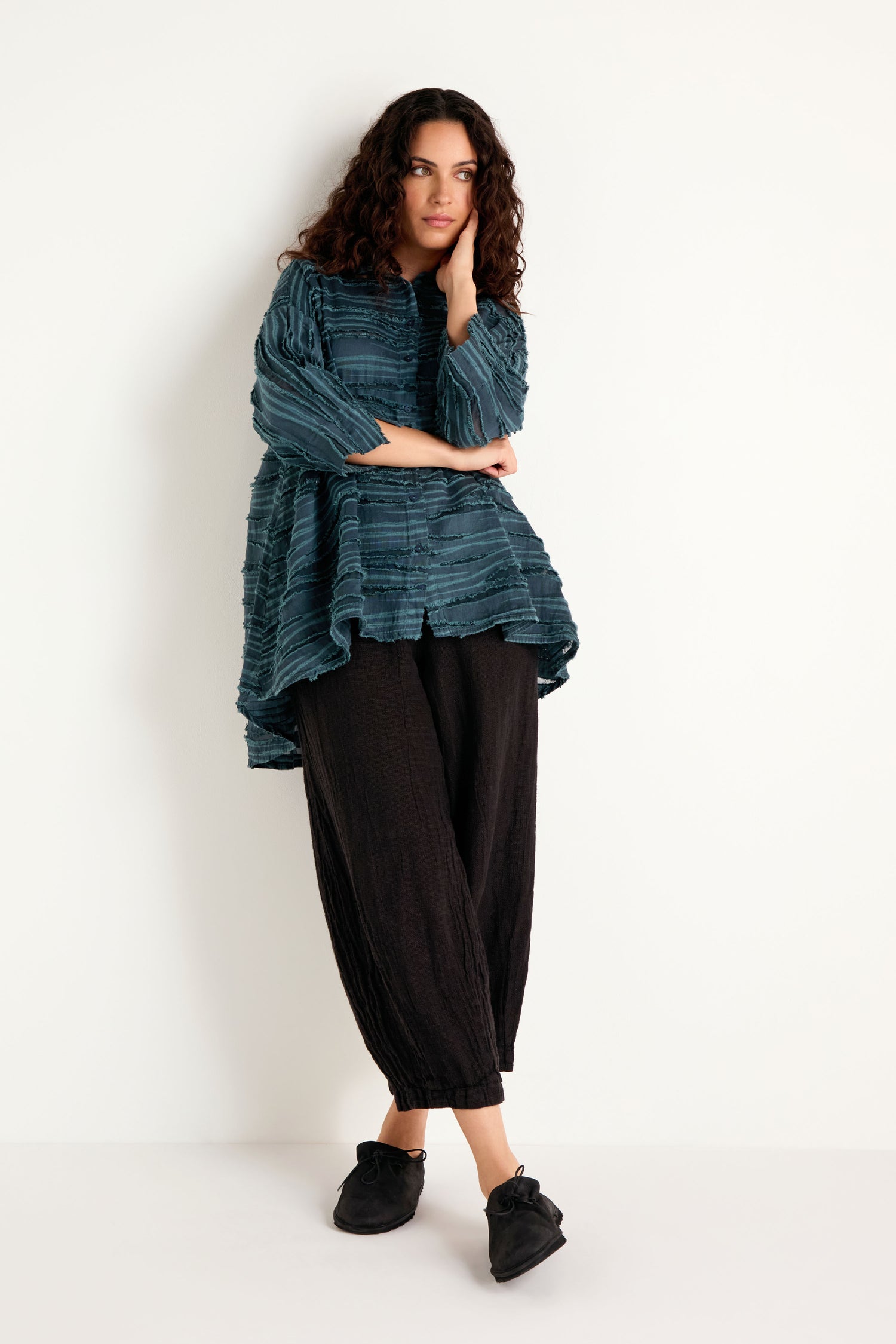 A woman with curly hair stands against a white wall wearing the Deep Turquoise Textured Stripe Flared Shirt, paired with black wide-leg pants and black shoes, with one hand touching her face.