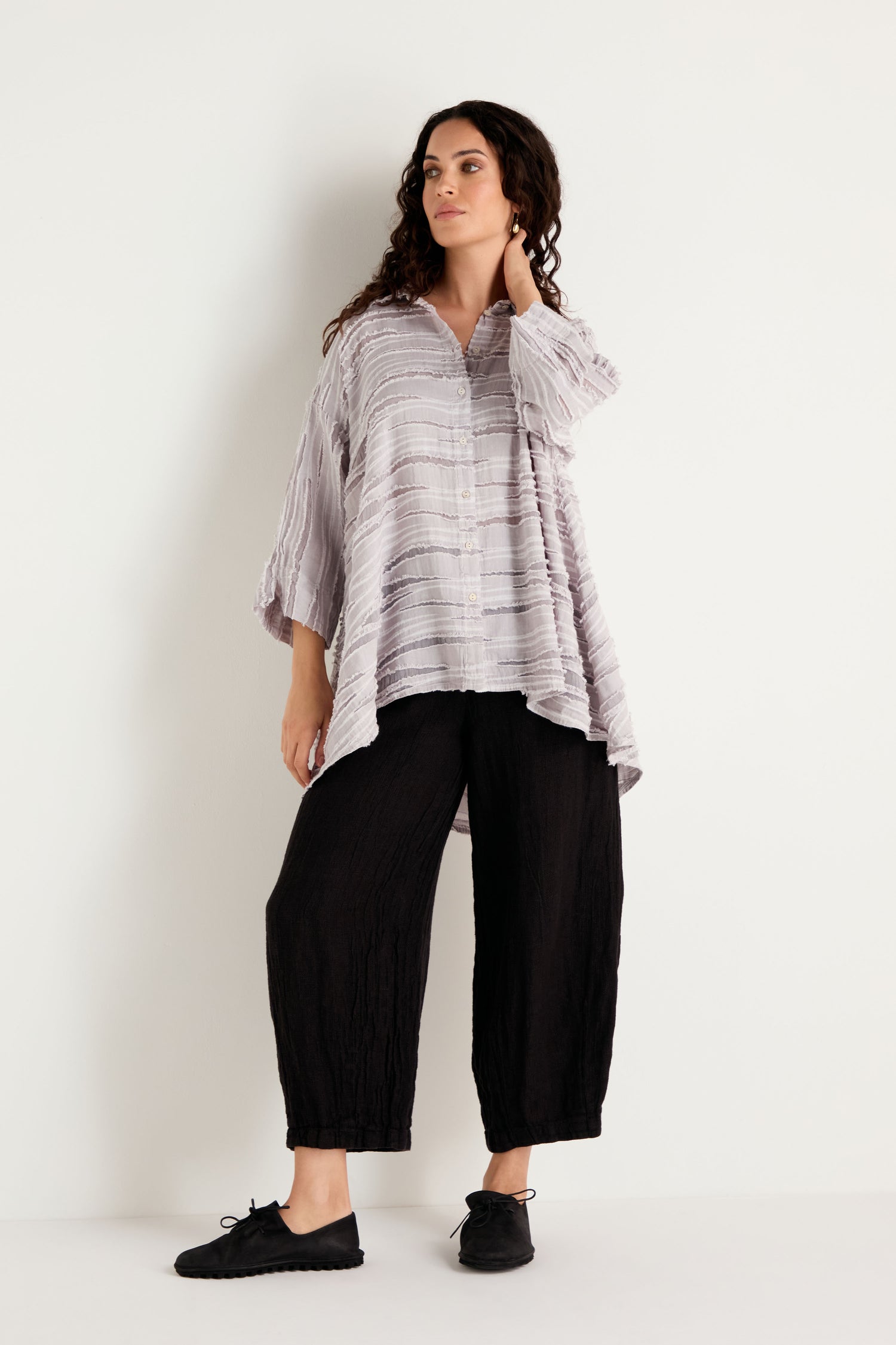 A woman with long curly hair stands against a white wall, her left hand touching her neck, wearing the Pearl Textured Stripe Flared Shirt in a loose, light gray linen silk cotton blend. She pairs it with black wide-leg pants and black shoes.