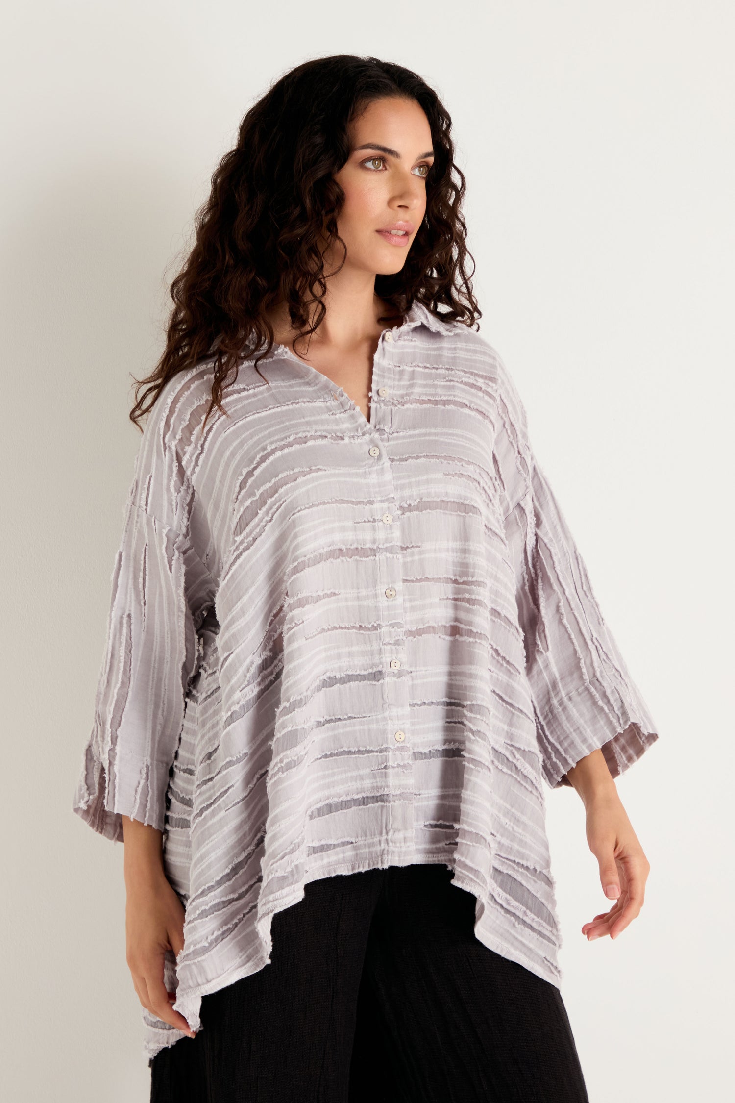 A woman with long curly hair wears the Pearl Textured Stripe Flared Shirt, a loose, light-colored top crafted from a linen silk cotton blend with an asymmetrical hem and textured detail, paired with black pants as she stands against a plain light background.