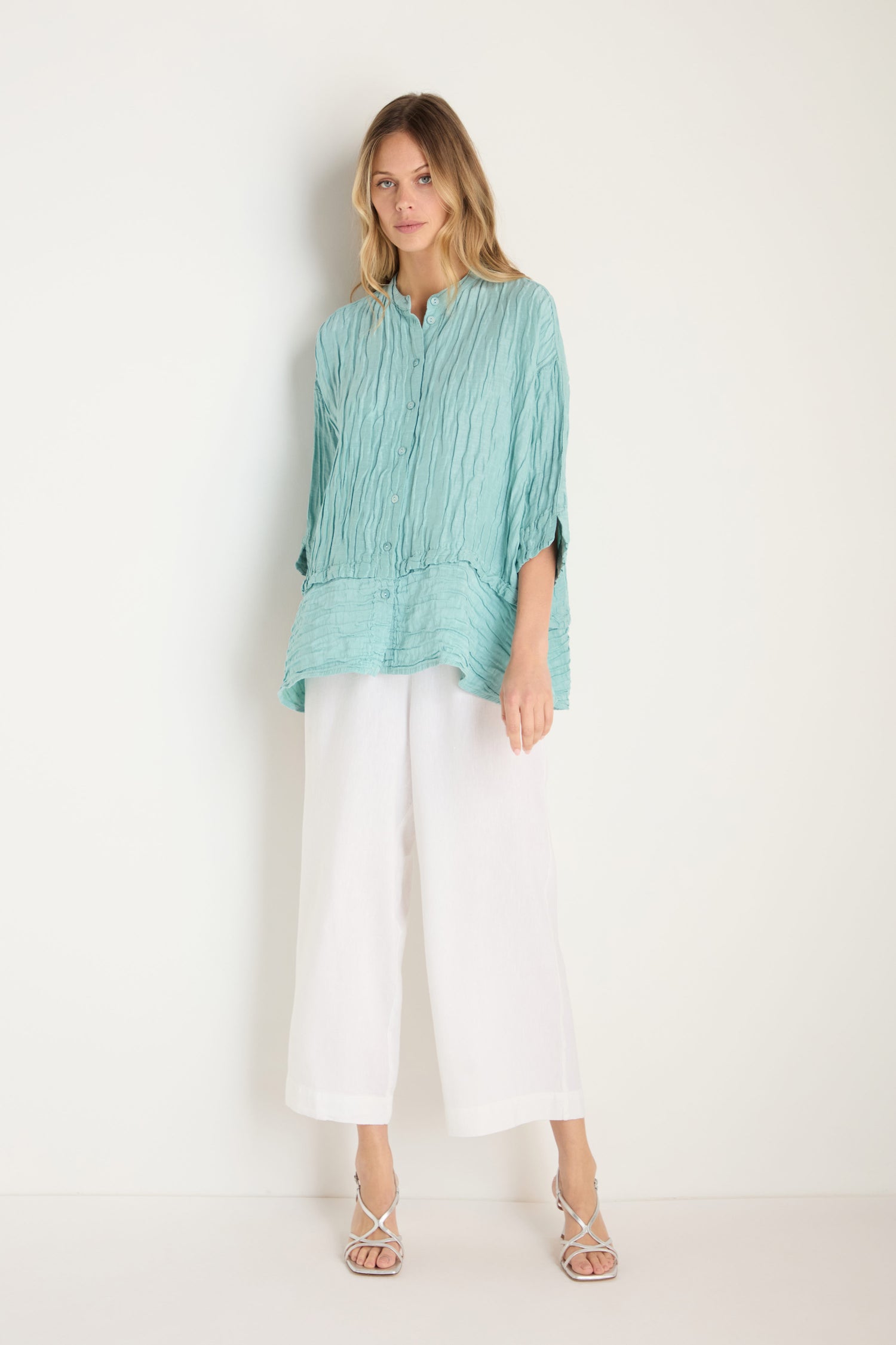 A woman stands against a white wall wearing the Panel Hem Silk Linen Shirt in light turquoise, featuring a breathable, oversized fit, paired with wide white pants and strappy white sandals.