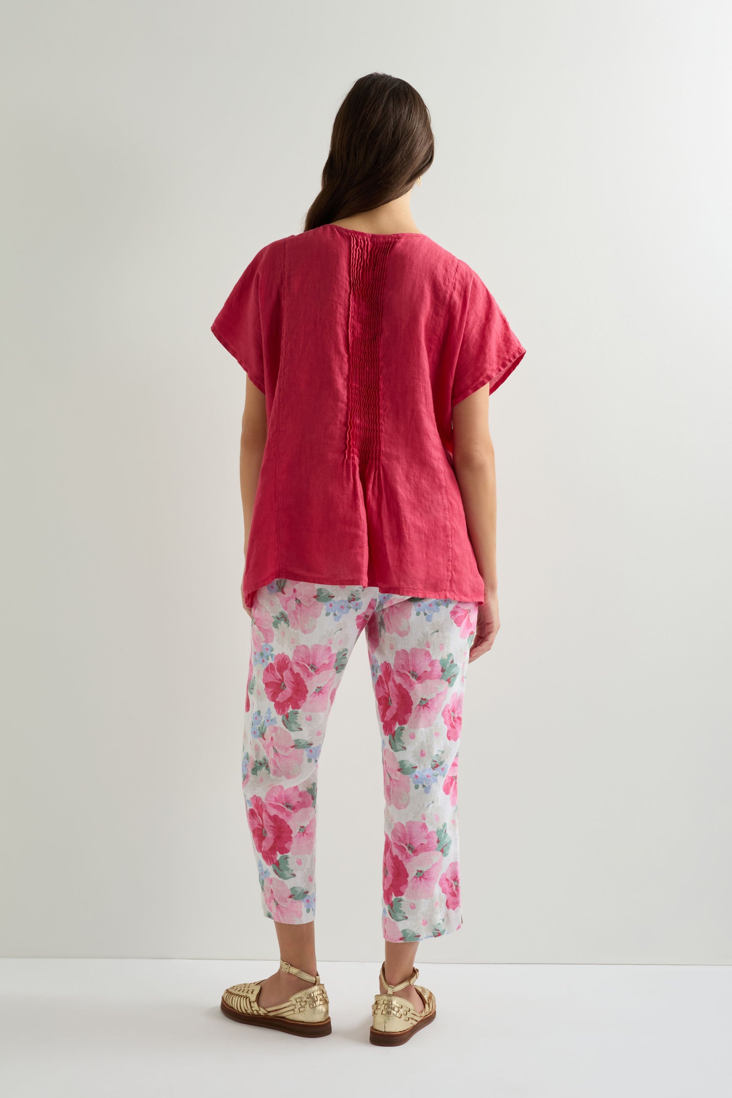 A woman faces away, wearing the Pleat Front Linen Top in pink with short sleeves, floral print pants, and tan sandals against a plain light background.