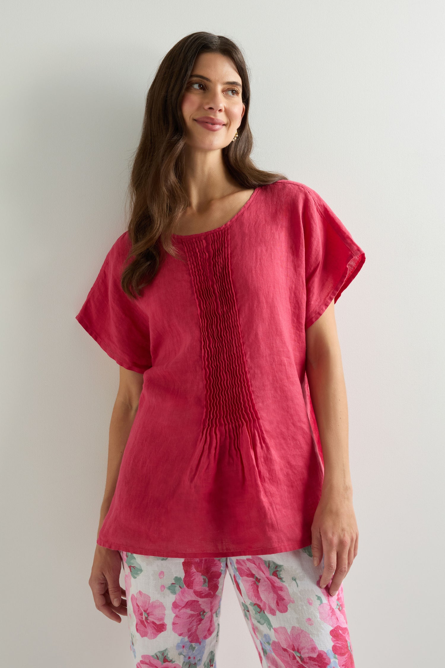 A woman stands against a light wall wearing the Pleat Front Linen Top in red, paired with white pants featuring large pink floral prints.
