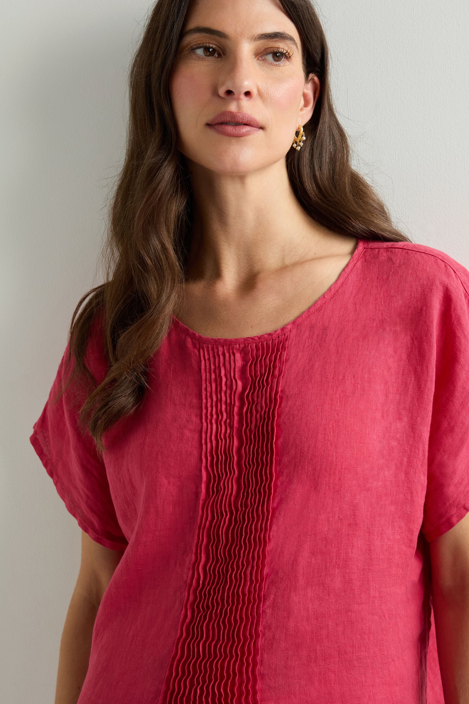 A woman with long brown hair wears the Pleat Front Linen Top in pink, made from breathable linen, as she stands against a light-colored wall.