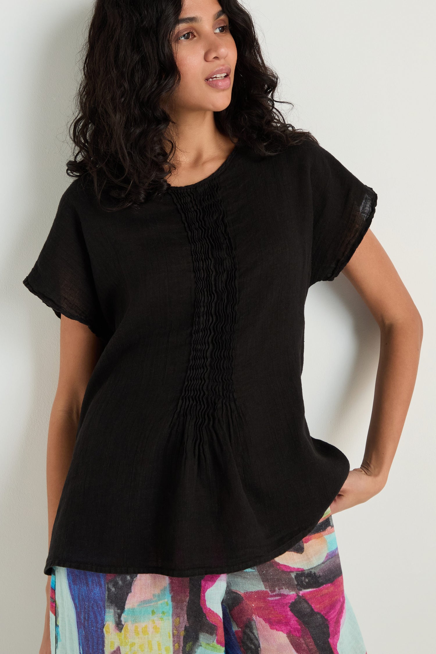 A woman wearing the Pleat Front Linen Top in black with pleat detailing and colorful abstract-patterned pants poses against a light background, highlighting the top's relaxed silhouette.