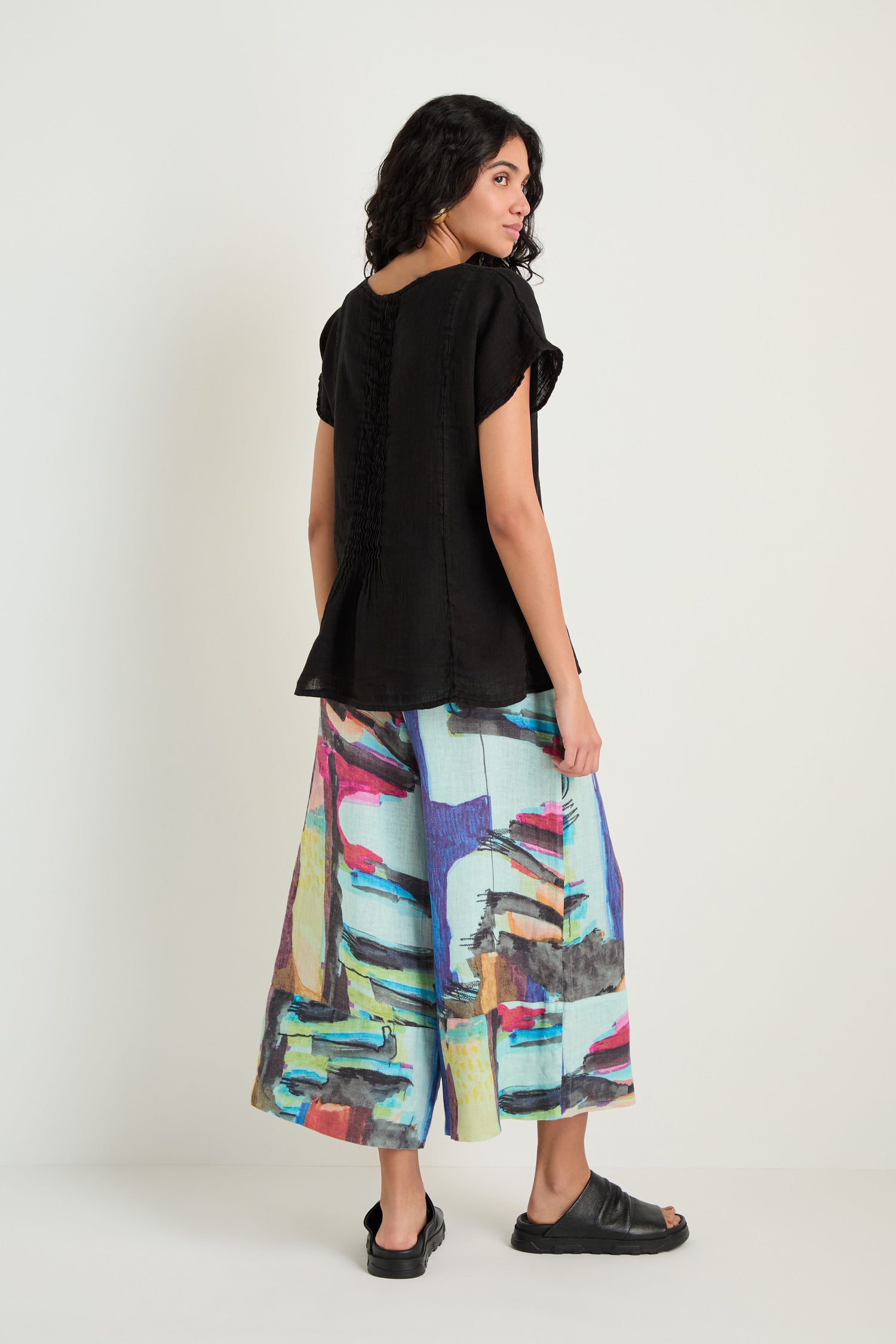 A woman stands against a plain background wearing the Pleat Front Linen Top in black and wide-leg pants with a colorful abstract pattern, paired with black sandals. She is turned slightly to the side, showcasing her outfit’s relaxed silhouette.