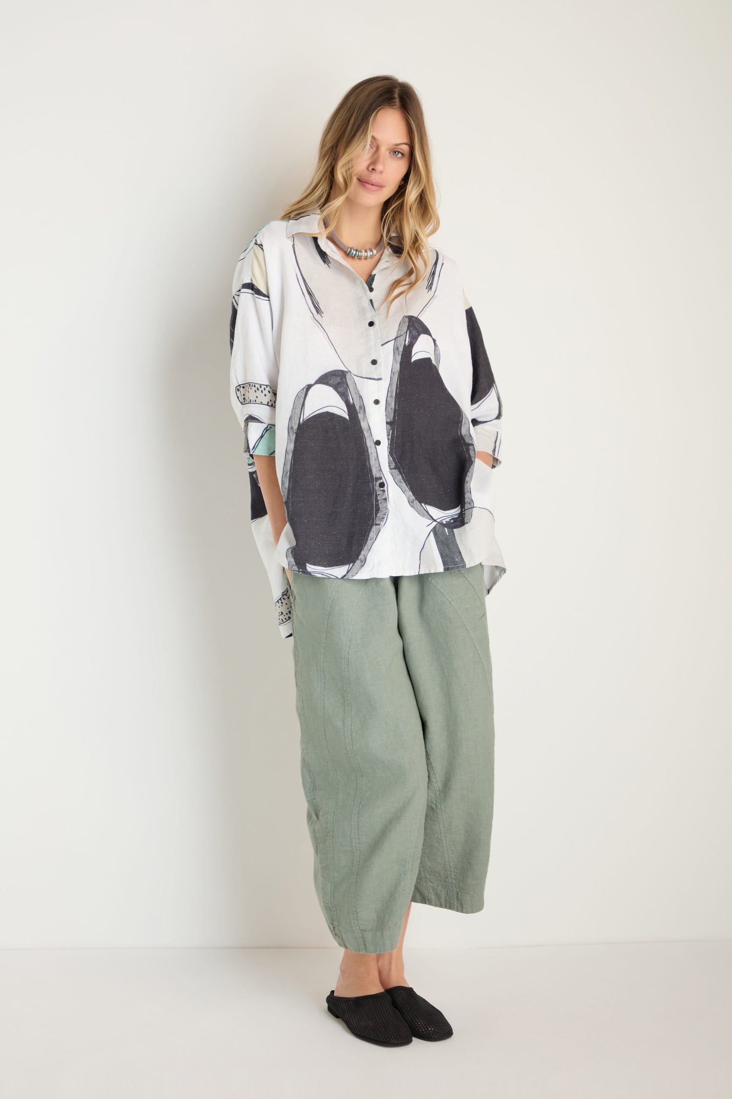 A woman stands against a plain wall wearing a Surreal Collage Print Linen Shirt, light green wide-leg pants, and black slip-on shoes.