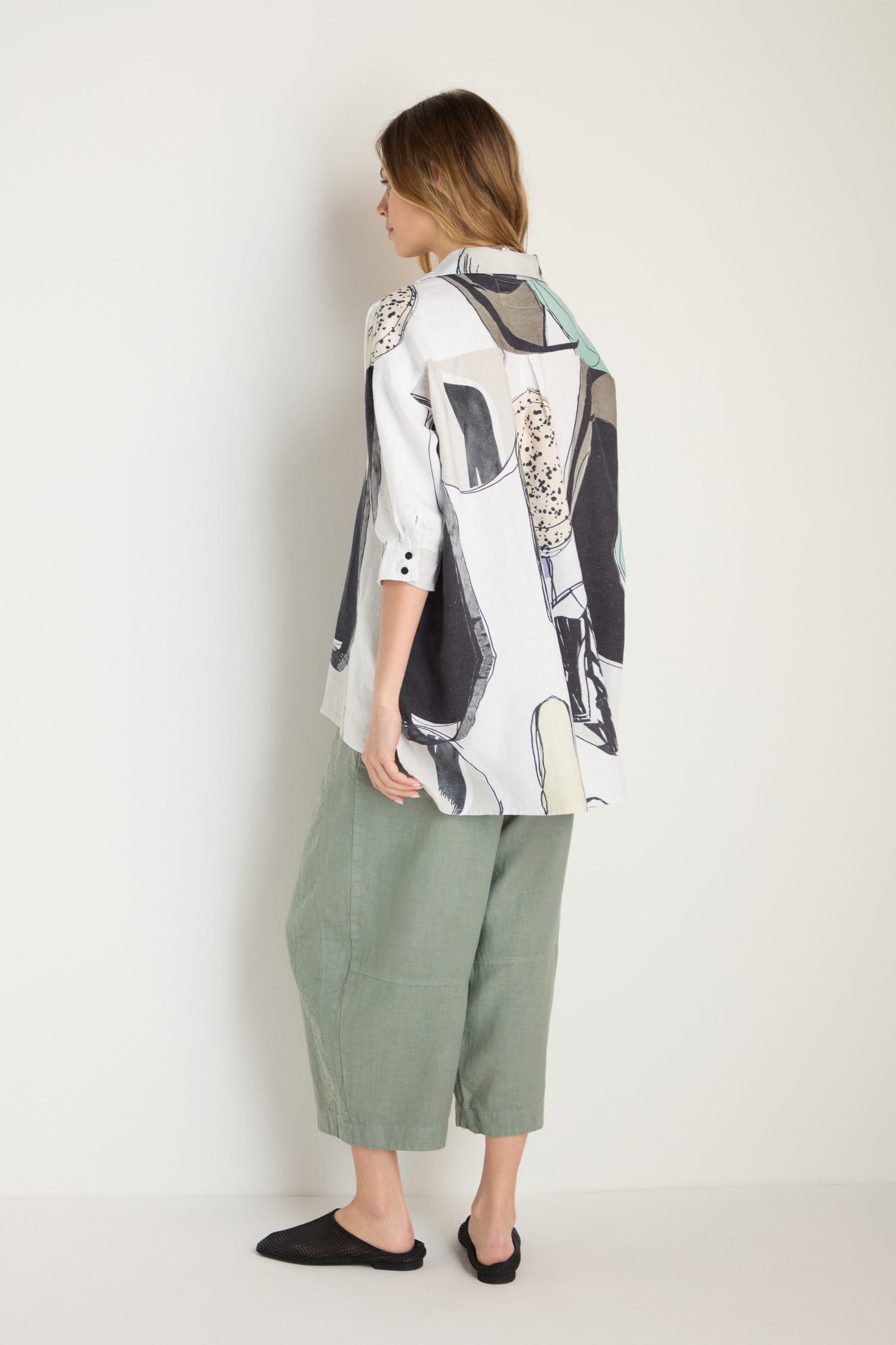 A woman faces away, wearing the Surreal Collage Print Linen Shirt, light green wide-leg pants, and black slip-on shoes against a plain background.