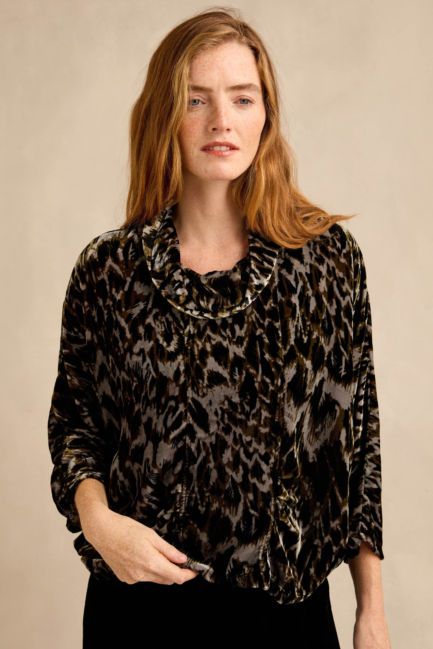 A woman with long red hair stands against a beige background wearing the Animal Print Cowl Neck Velvet Top, a loose-fitting black and brown velvet piece.