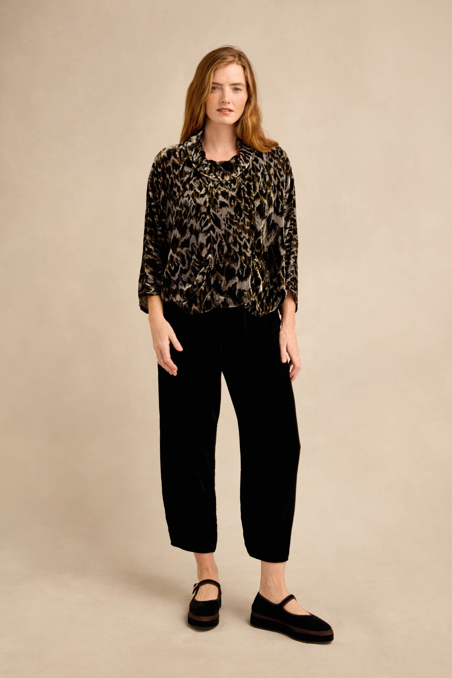 A person stands against a neutral background wearing the Animal Print Cowl Neck Velvet Top, paired with black wide-leg pants and black Mary Jane shoes.