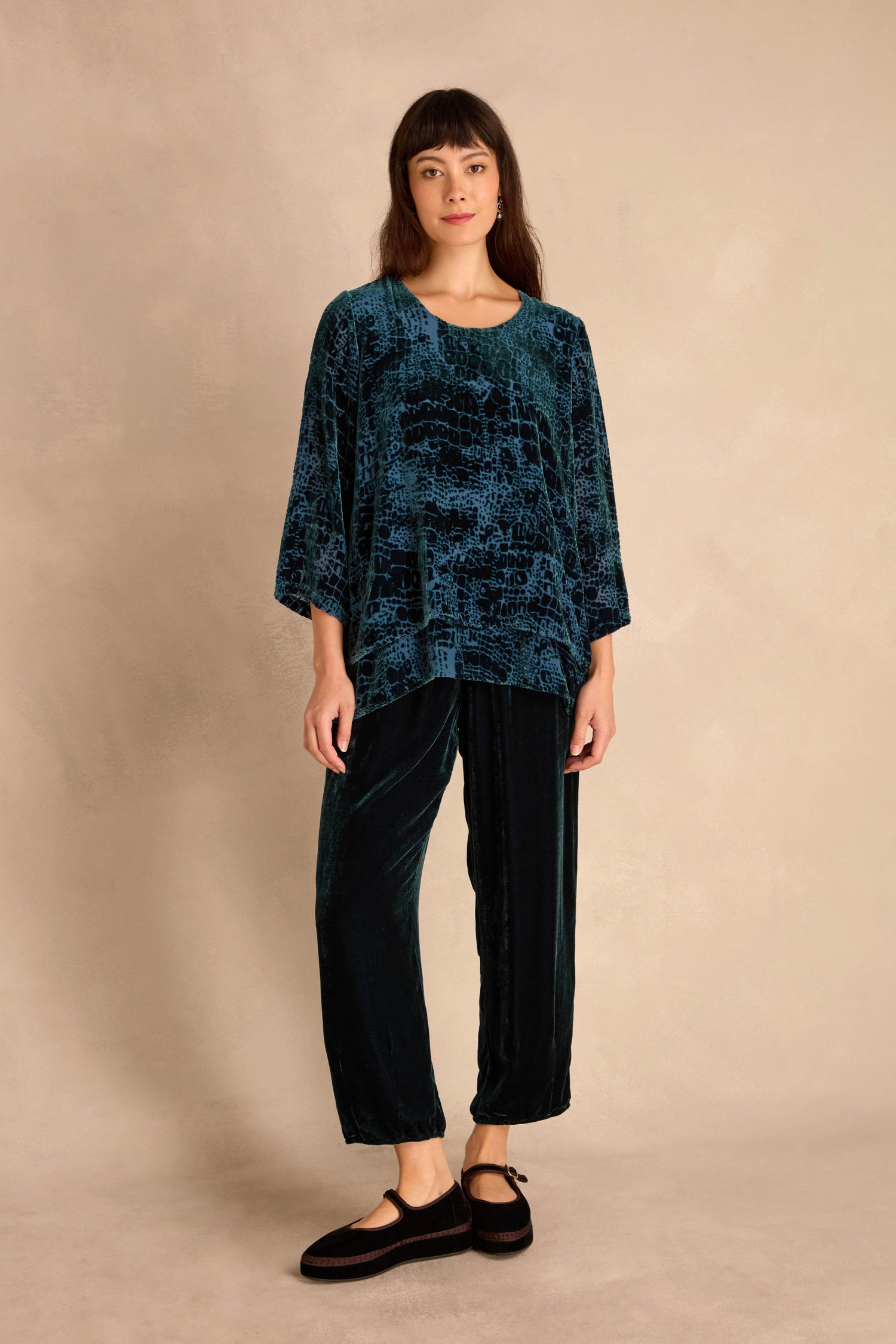 A person stands against a beige background wearing the Deep Turquoise Leopard Devore Layered top, matching relaxed pants, and black flat shoes.