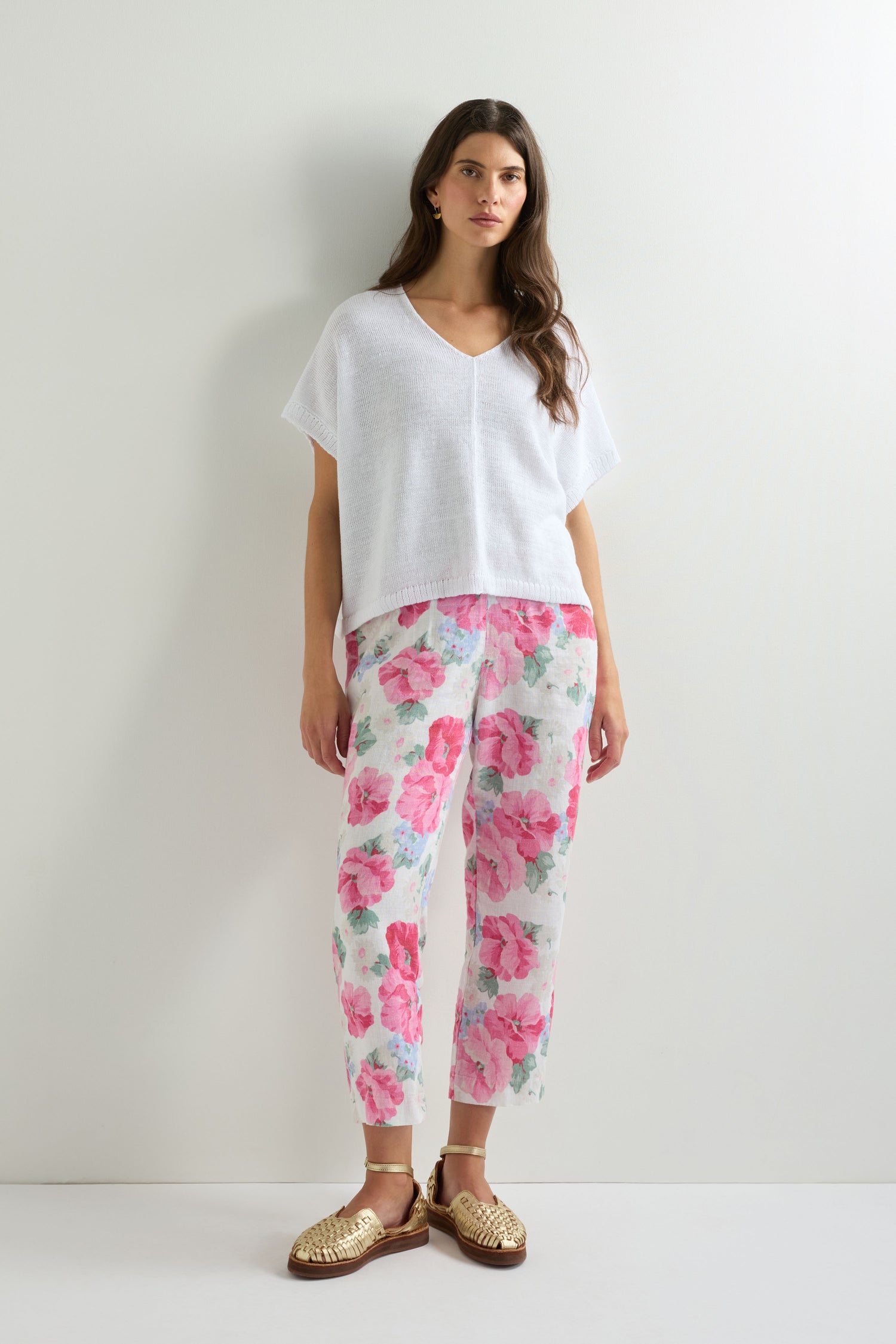 A woman stands against a white wall, wearing the Sleeveless V Neck Boxy Linen Knit top, floral pink and white pants, and gold woven shoes.