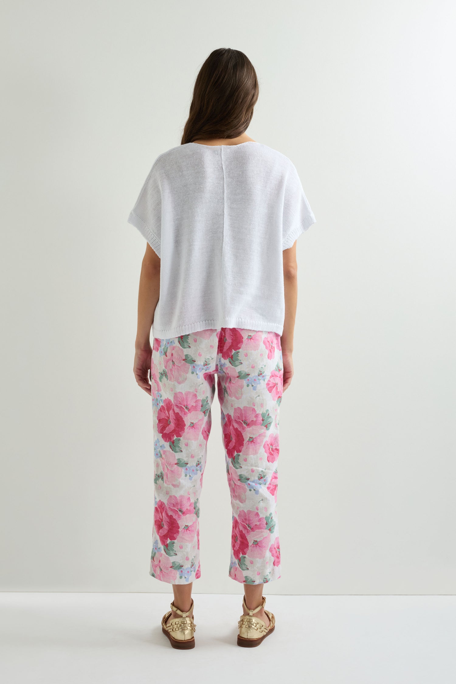A woman faces away in a Sleeveless V Neck Boxy Linen Knit top, paired with pink floral pants and gold artisanal sandals, set against a plain white background.
