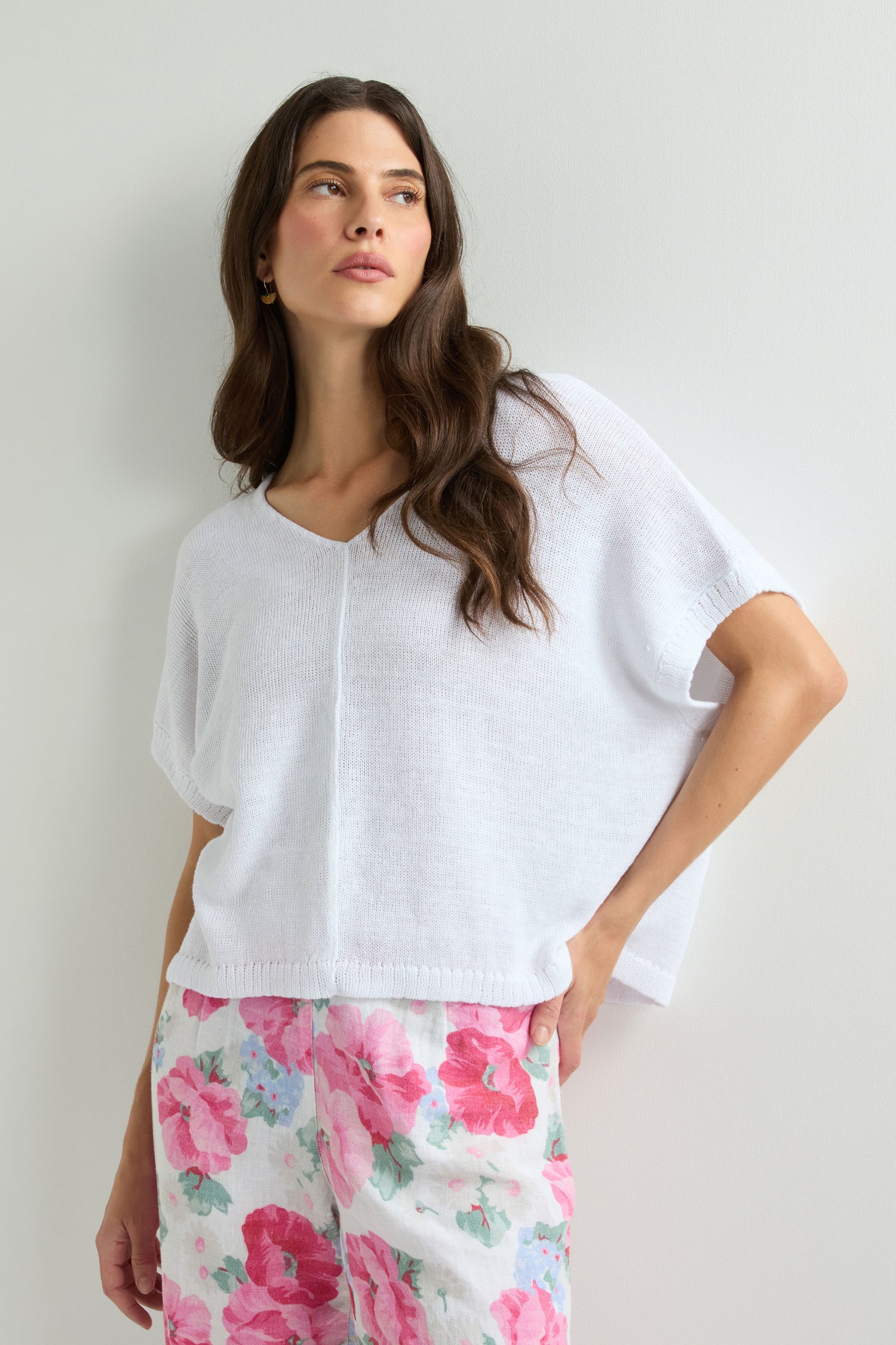 A woman wearing the Sleeveless V Neck Boxy Linen Knit stands against a plain light-colored wall, paired with floral print pants and looking to the side.