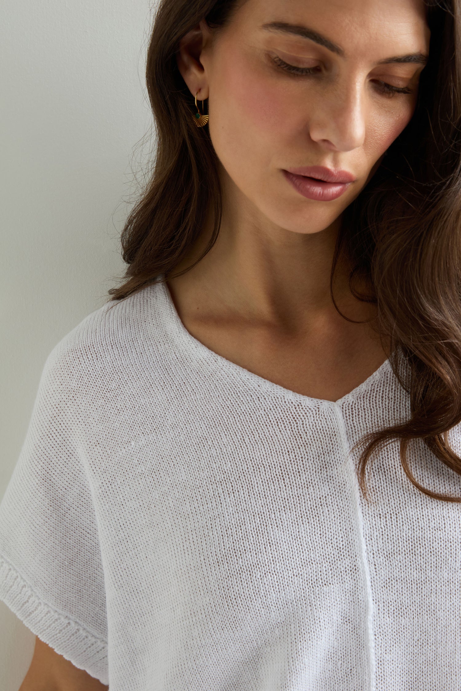 Woman with long brown hair wears the Sleeveless V Neck Boxy Linen Knit layered under a linen vest, paired with gold hoop earrings. She looks down with a neutral expression.