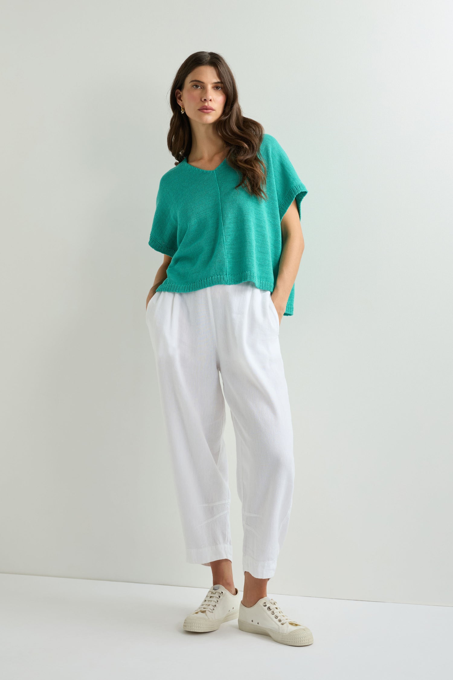 A woman stands against a white wall wearing a loose teal top under the Sleeveless V Neck Boxy Linen Knit vest, paired with white pants and sneakers, her hands in her pockets.