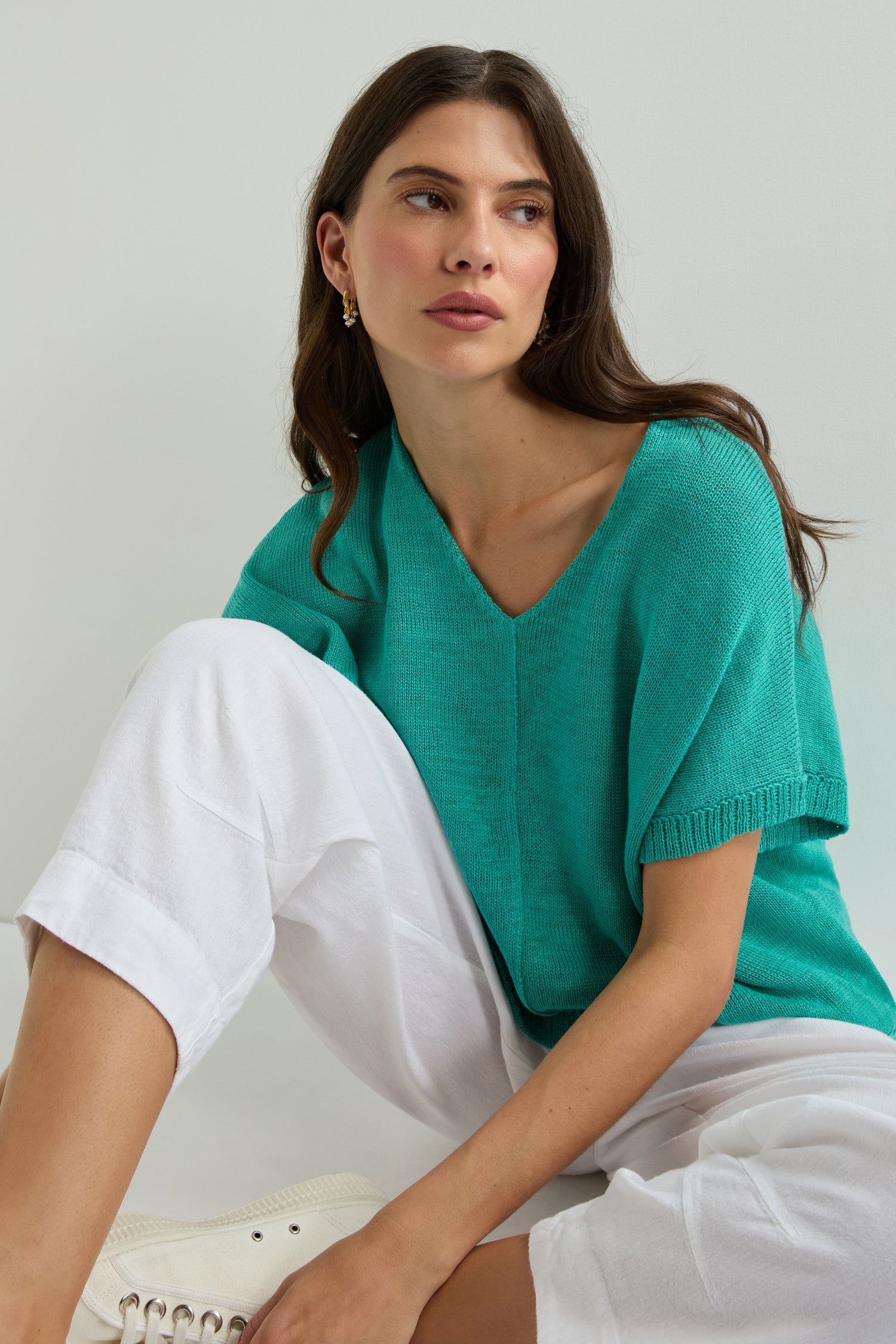 A woman with long brown hair wears the Sleeveless V Neck Boxy Linen Knit in teal, paired with white pants and gold hoop earrings, sitting against a plain light background.