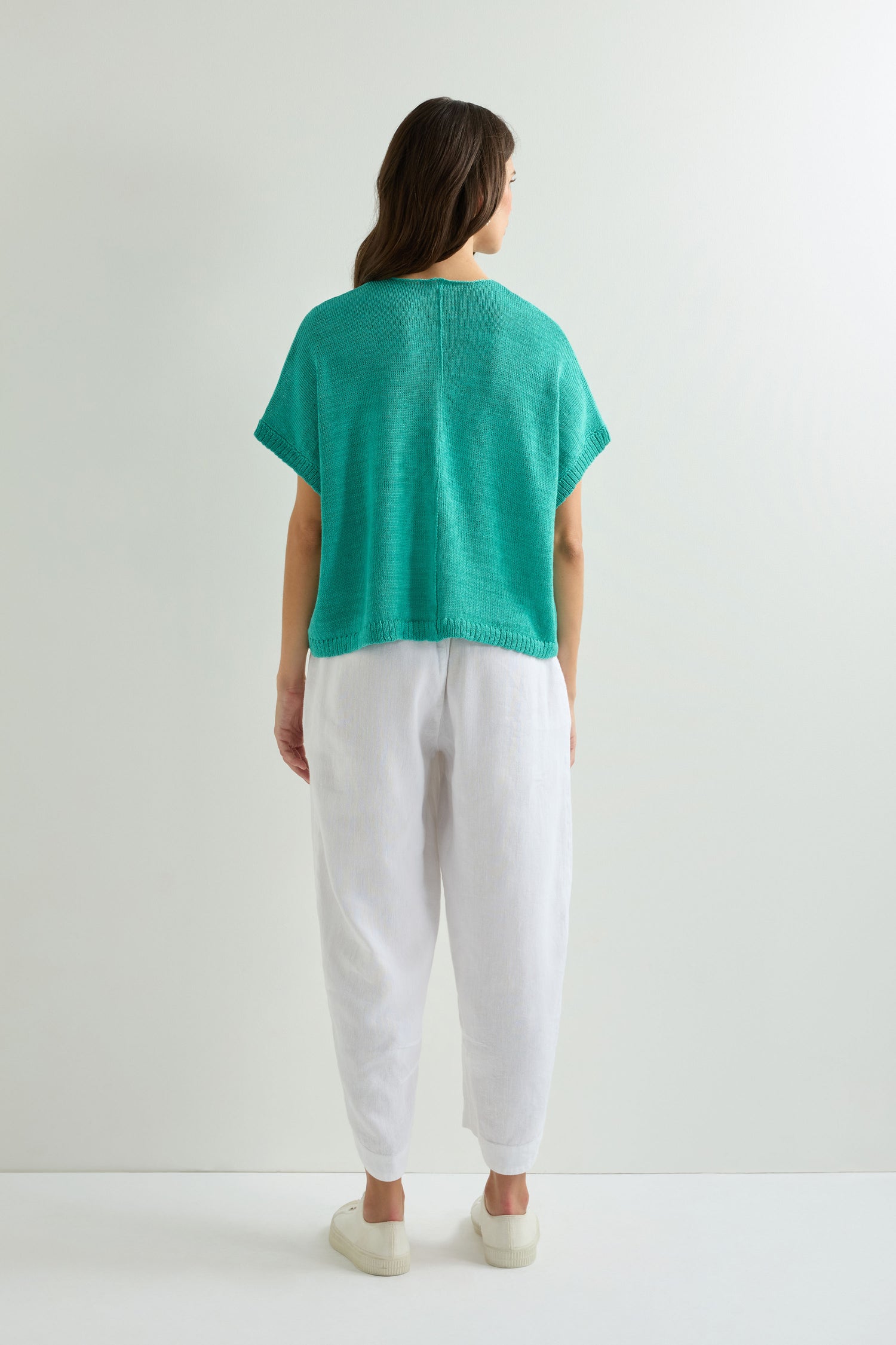 Person stands against a plain white wall, wearing the Sleeveless V Neck Boxy Linen Knit in turquoise, paired with white pants and shoes; shown from the back.