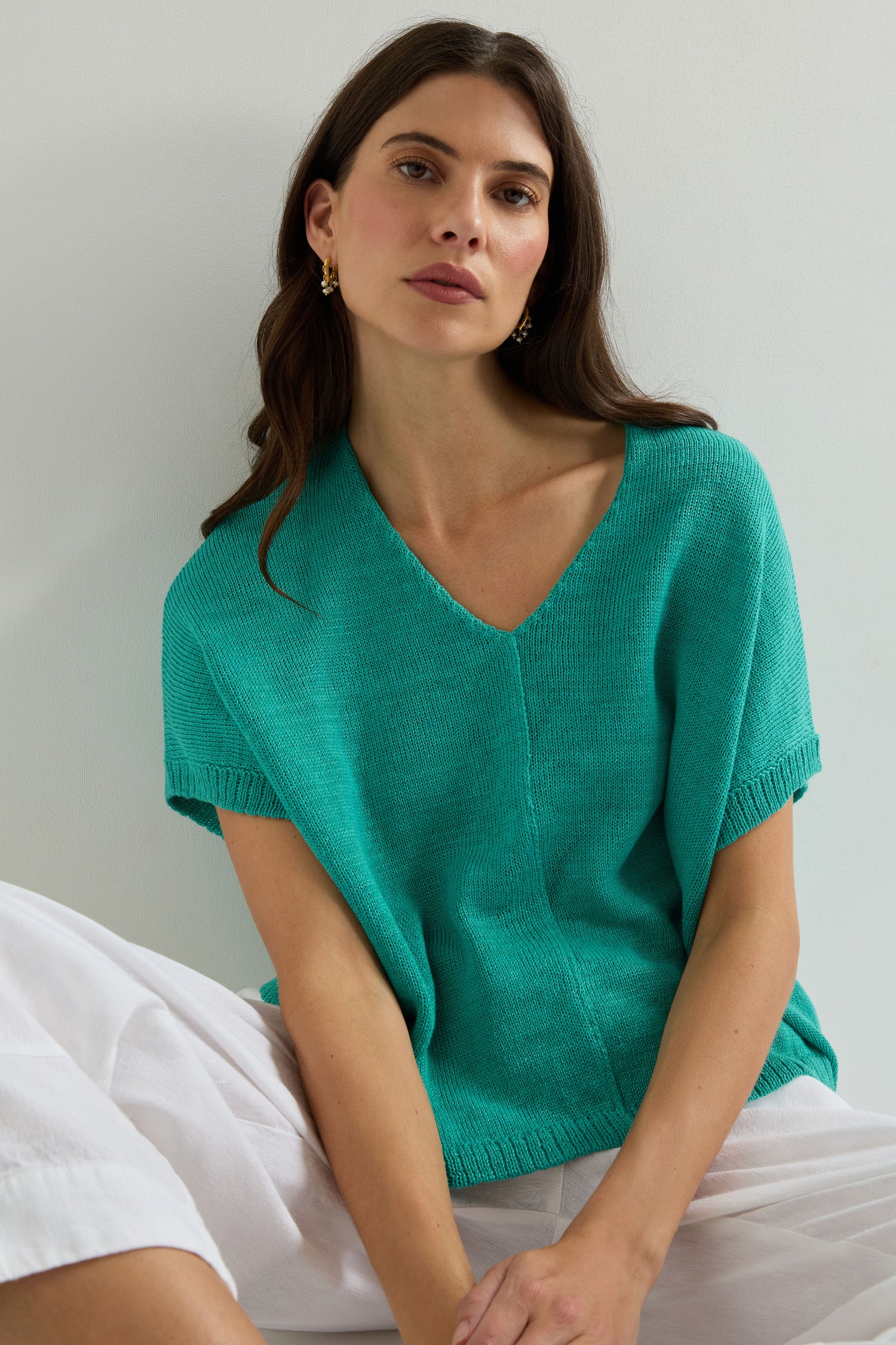 A woman with long brown hair wears the Sleeveless V Neck Boxy Linen Knit over a loose turquoise top and white pants, sitting against a light wall.