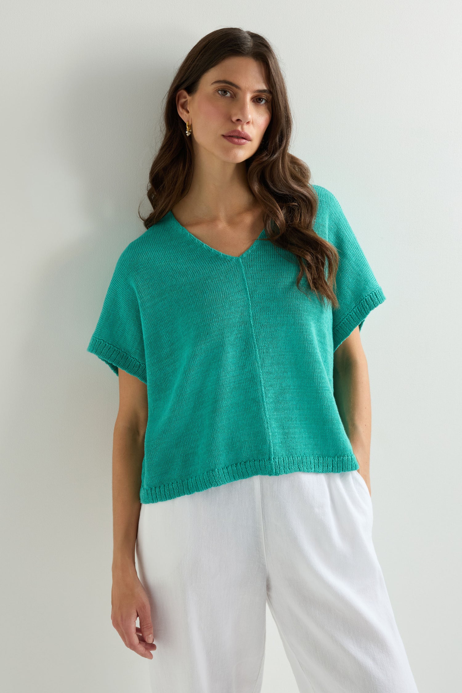 A woman stands against a white wall wearing a turquoise Sleeveless V Neck Boxy Linen Knit top with an artisanal finish and white pants.