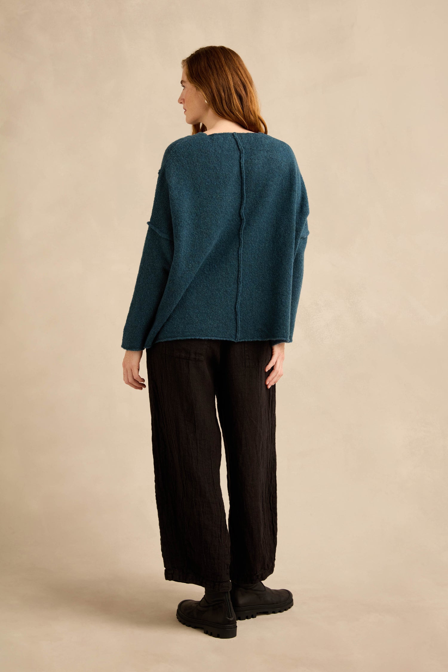 A person stands facing away, wearing the Deep Turquoise Seamed Knit with subtle curved seam detailing, paired with loose black pants and black shoes, against a plain beige background.
