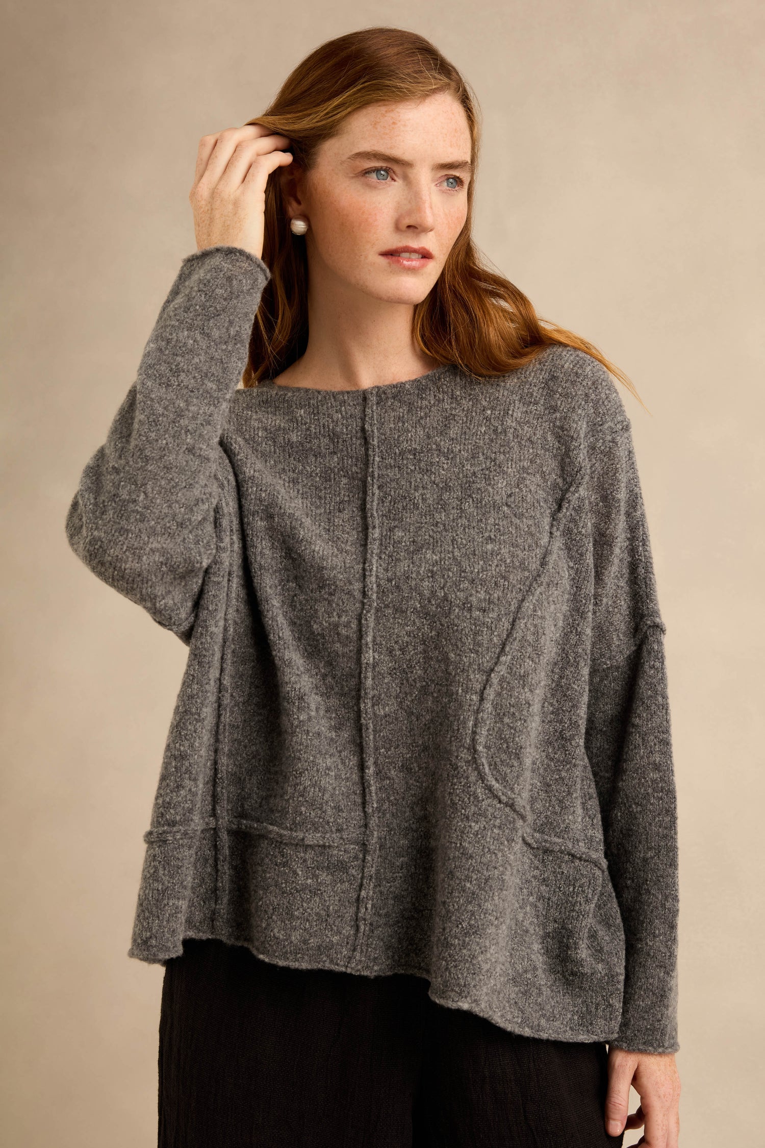 A woman stands against a neutral background, looking to the side with one hand touching her hair, wearing the Grey Seamed Knit sweater paired with black pants.