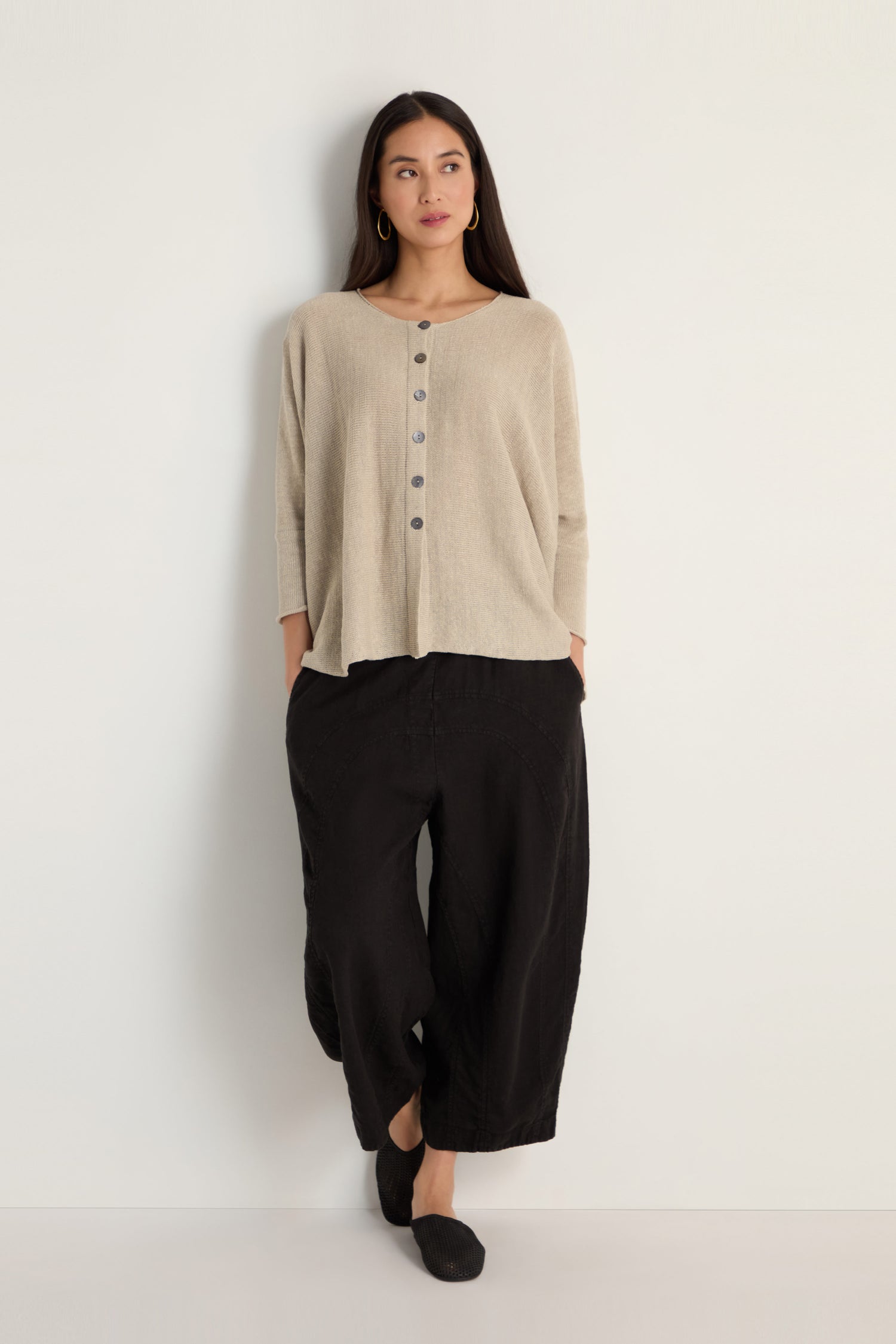 A woman stands against a white wall, wearing the beige Boxy Linen Button Cardigan, loose black pants, earrings, and black slip-on shoes.