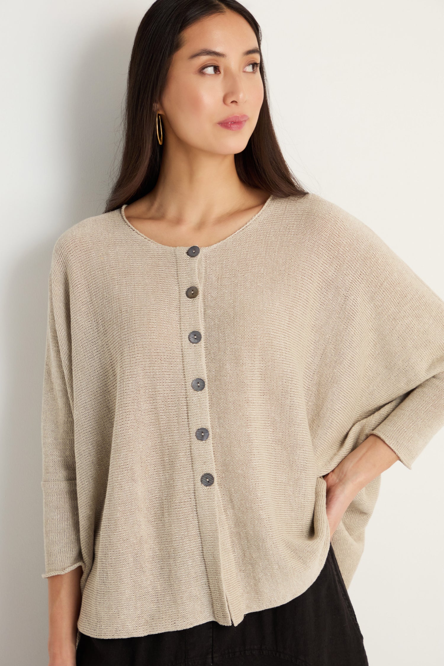 A woman with long brown hair wears the Boxy Linen Button Cardigan in beige against a plain background, looking to the side with one hand on her hip—a perfect lightweight layering piece.