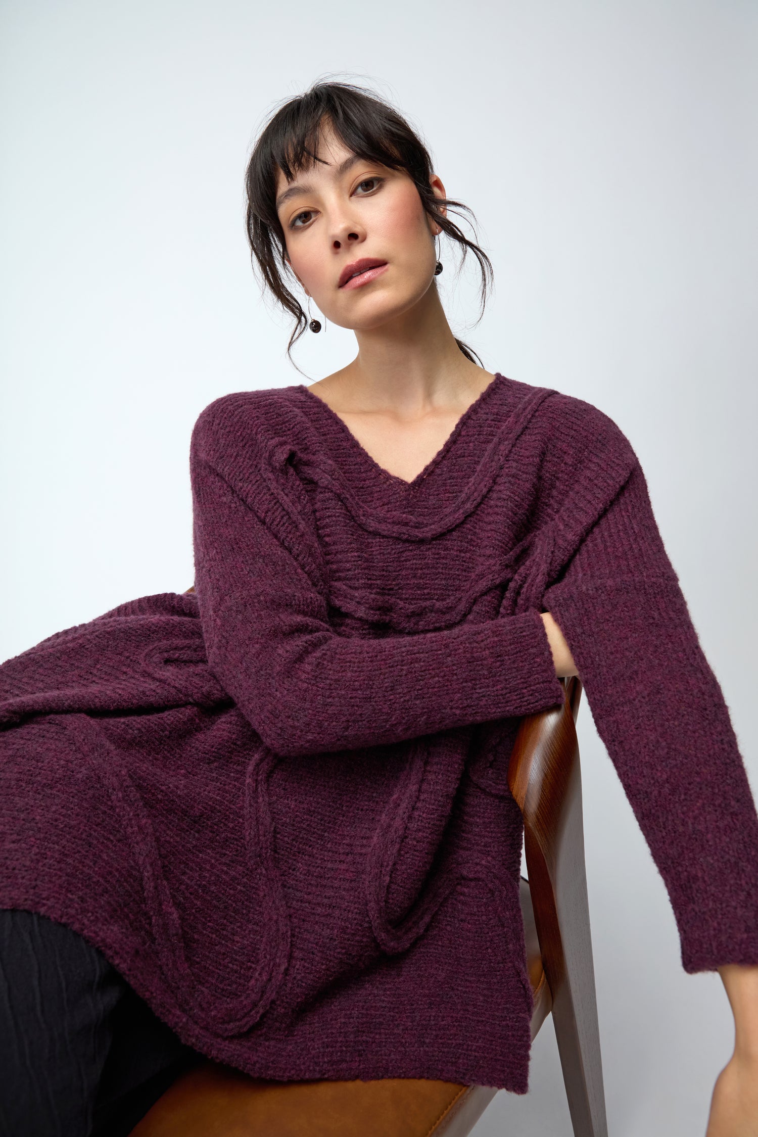 Woman sitting on a chair wearing the Swirl Seamed Oversize Aubergine Knit sweater with subtle seam detailing and black pants, set against a plain light background.