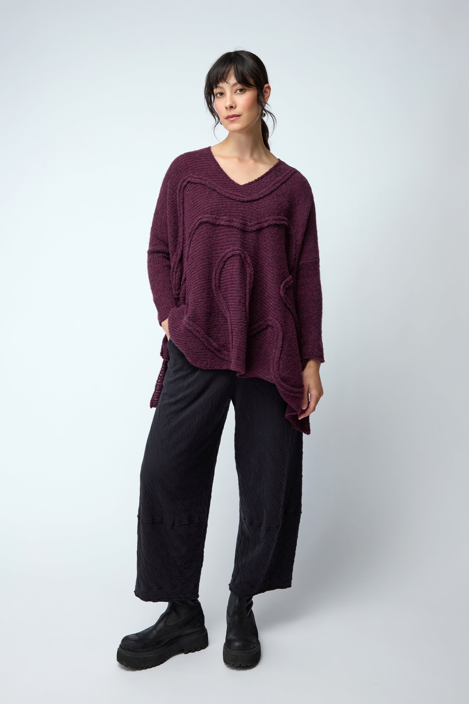 A woman stands against a light background wearing the Swirl Seamed Oversize Aubergine Knit, paired with wide black pants and black boots, her hands by her sides.
