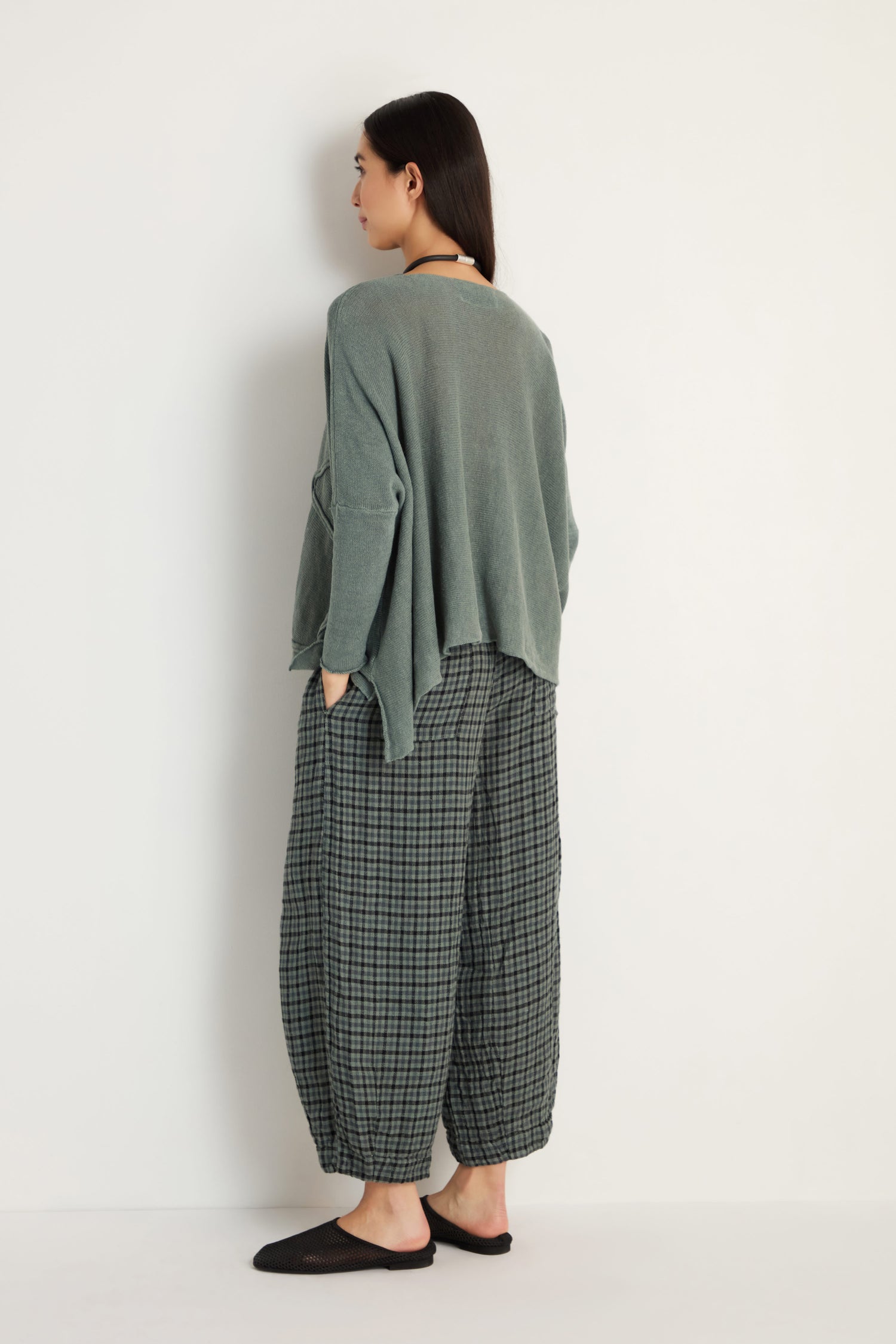 A person stands facing away, wearing the Swirl Seamed Linen Knit Top in sage green with a relaxed fit, paired with checkered gray-green wide pants and black slip-on shoes against a plain white background.