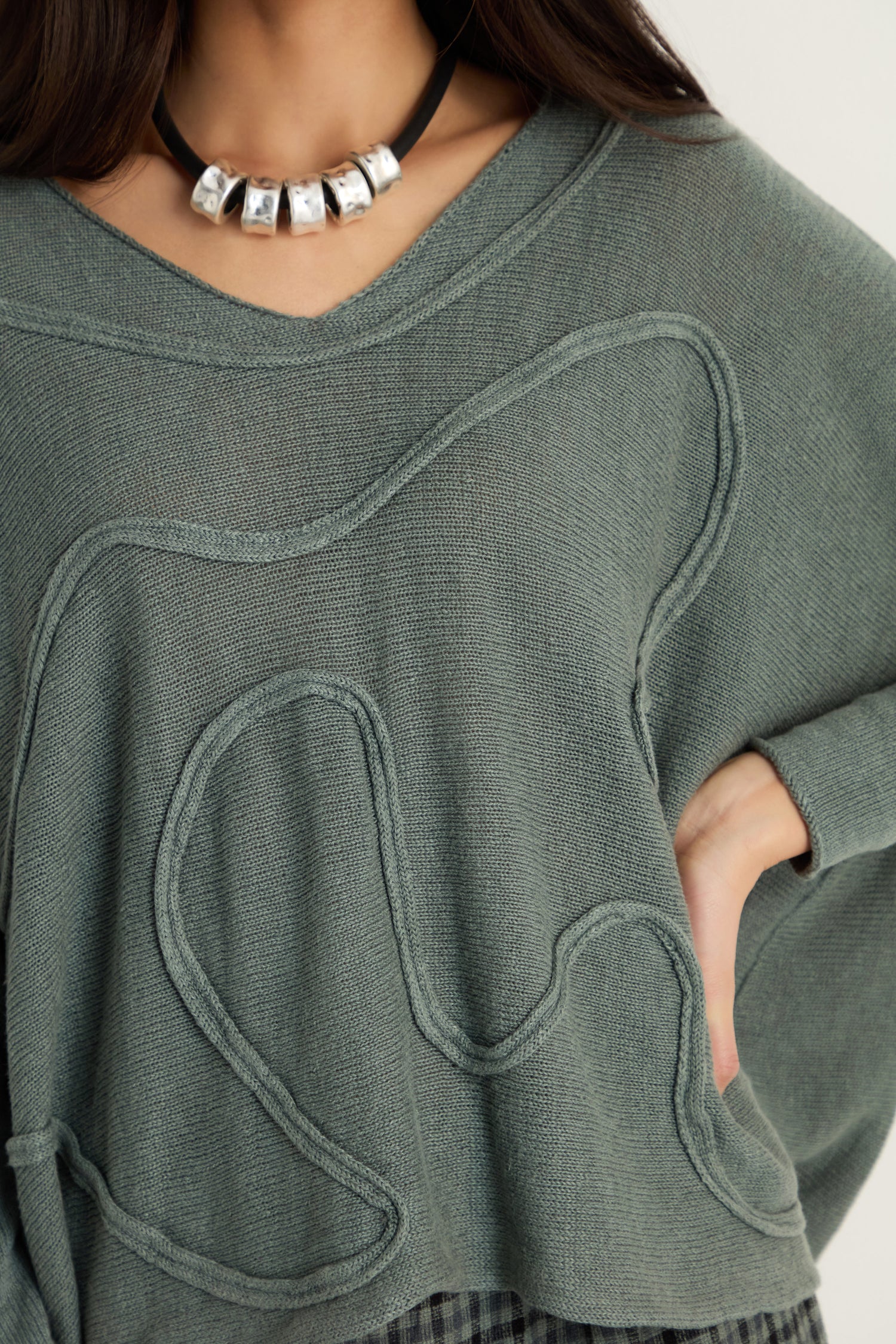 A person wears the Swirl Seamed Linen Knit Top, featuring green textured linen with swirl seam detailing and a relaxed silhouette, paired with a chunky silver bead necklace. Their hand on their hip highlights the casual style.