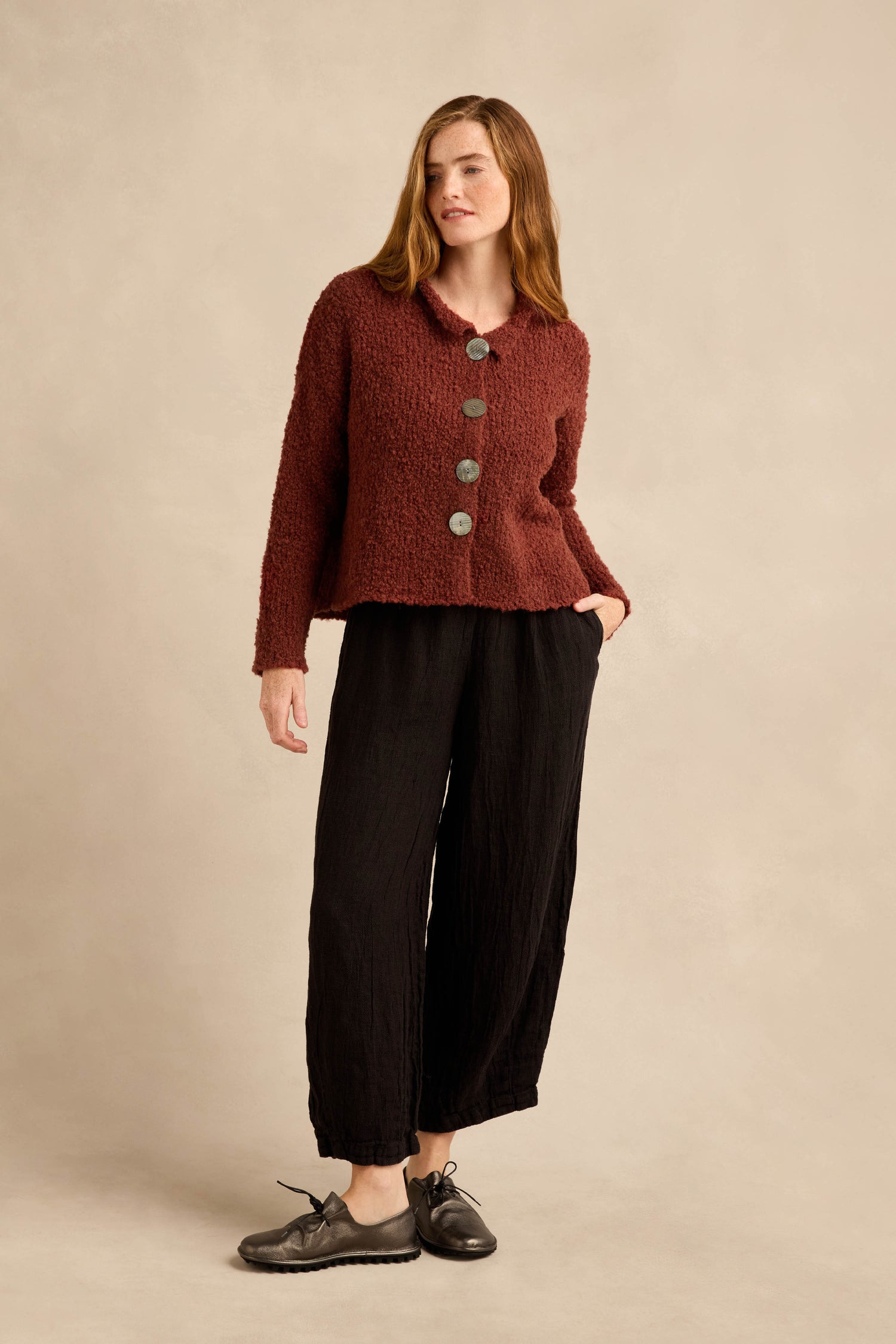 A woman stands against a beige background wearing the Three Button Cardigan, an oversized piece in textured burgundy knit, paired with loose black pants and black shoes, with one hand in her pocket.