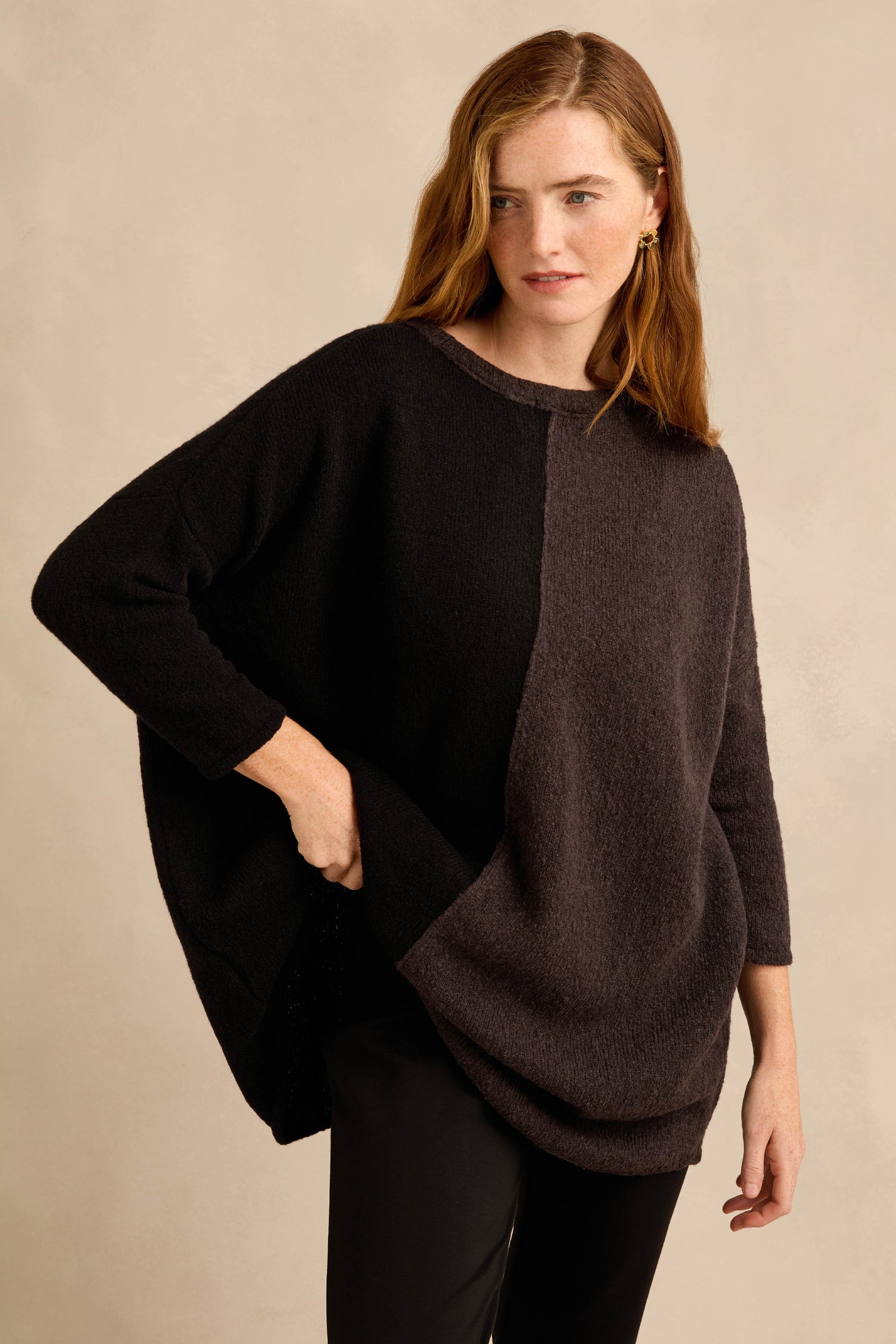 A woman is wearing the Two Colour Knit, a loose, long-sleeved sweater with an asymmetric colorblock design in black and dark gray, paired with black pants and standing against a neutral background.