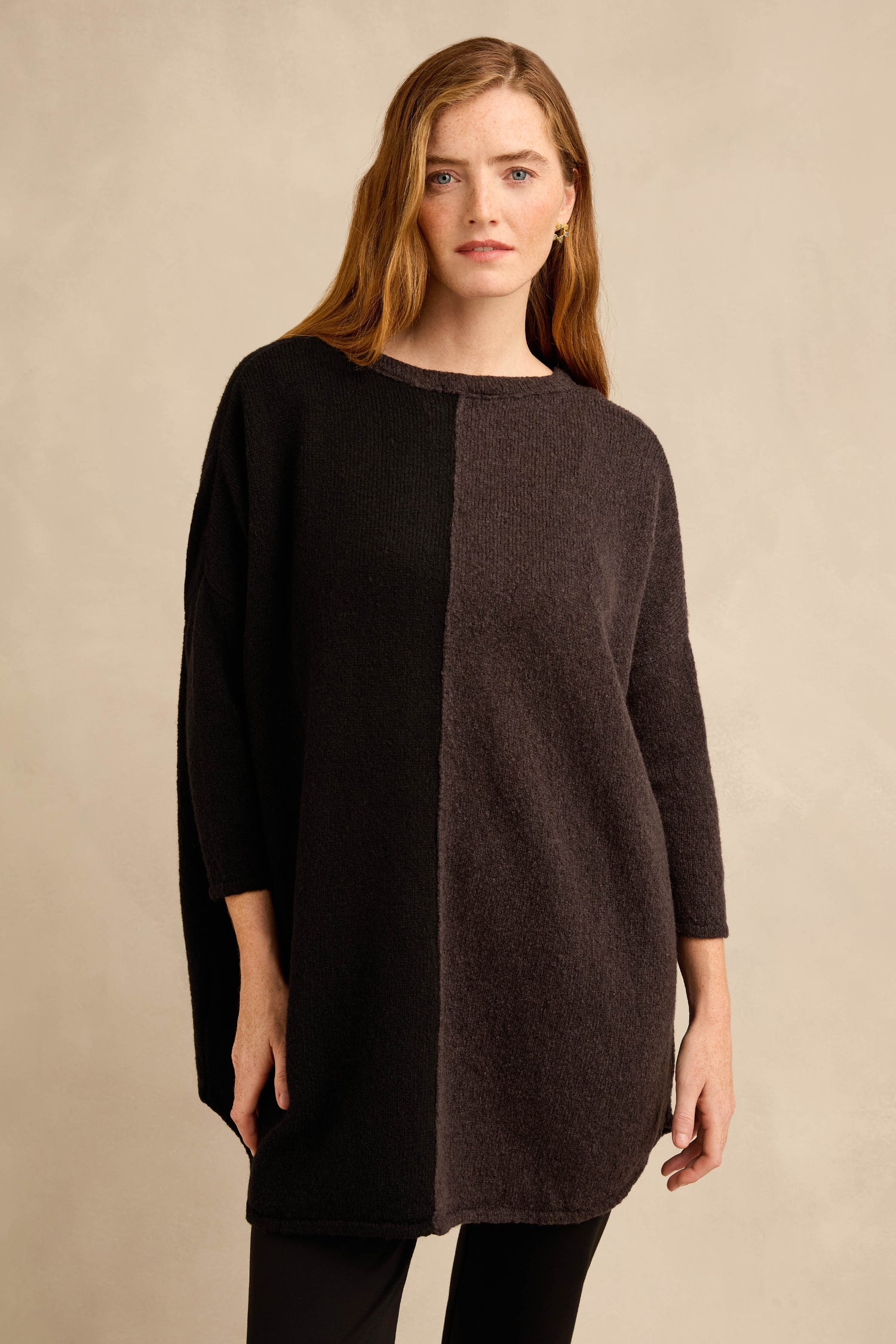 A woman wears the Two Colour Knit, a long-sleeved, oversized wool-rich tunic featuring a vertical color block design in dark gray and black and a relaxed silhouette, as she stands against a neutral background.
