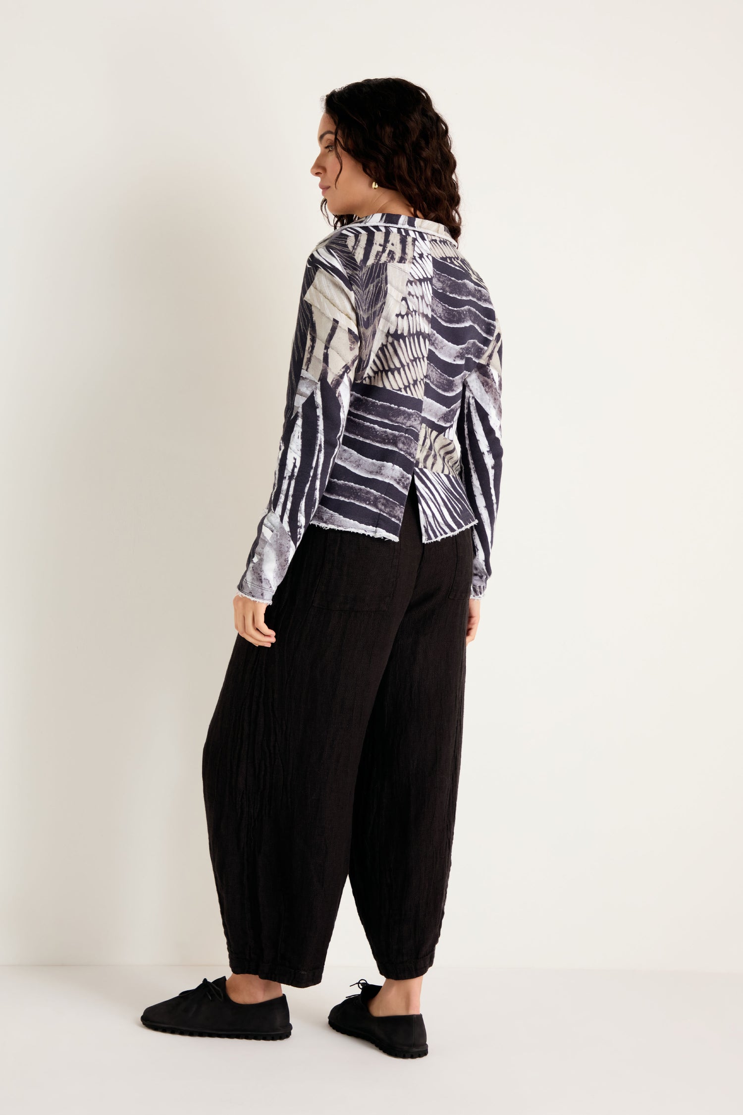 A person stands against a plain background, facing away from the camera, wearing a patterned long-sleeve shirt and loose black pants paired with black shoes; their outfit is enhanced by the Monochrome Patchwork Jersey Jacket, which adds an extra layer of texture.