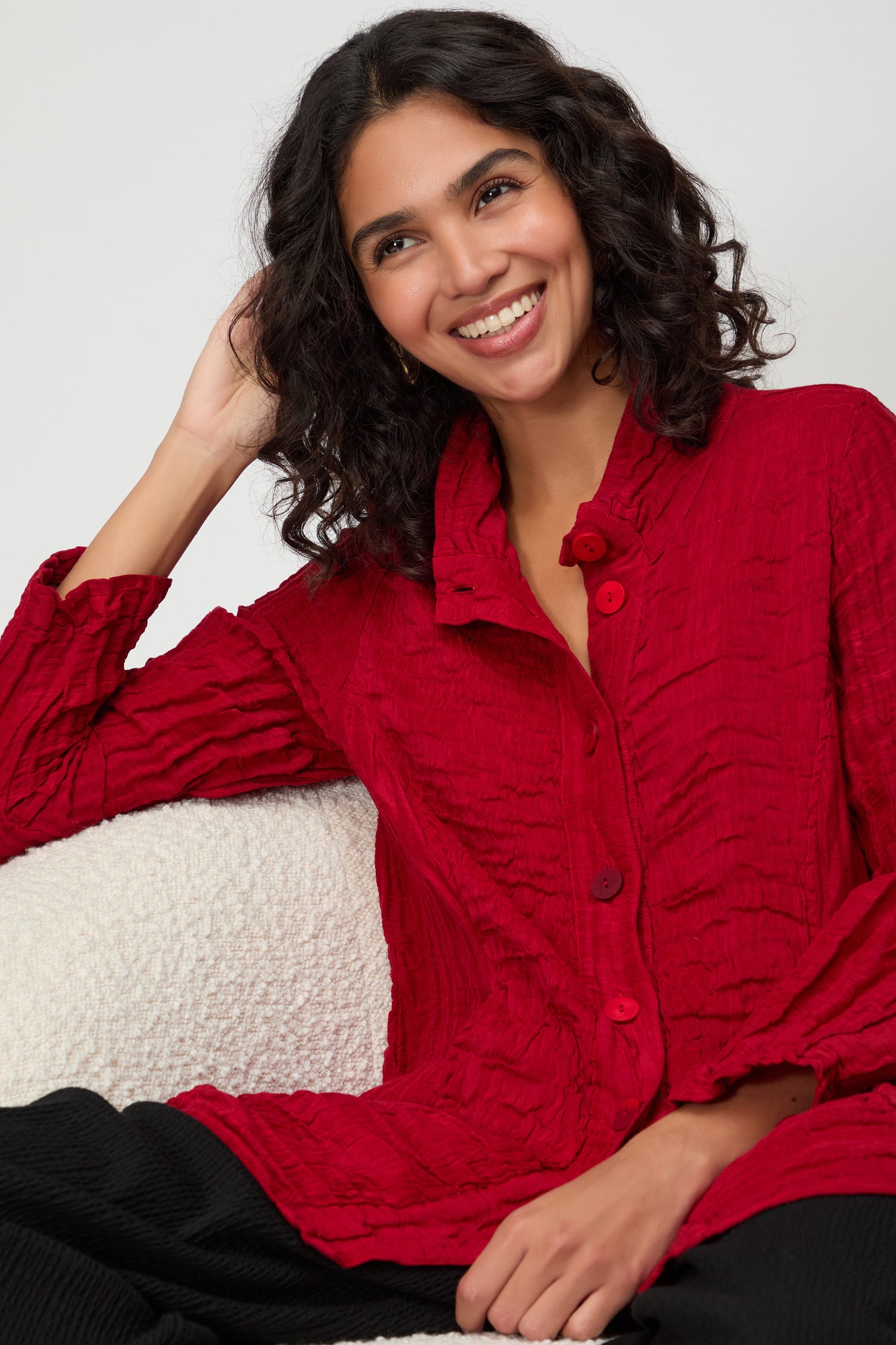 A woman with curly dark hair smiles while seated against a plain background, wearing the Panel Front Silk Linen Jacket in textured red and black pants.