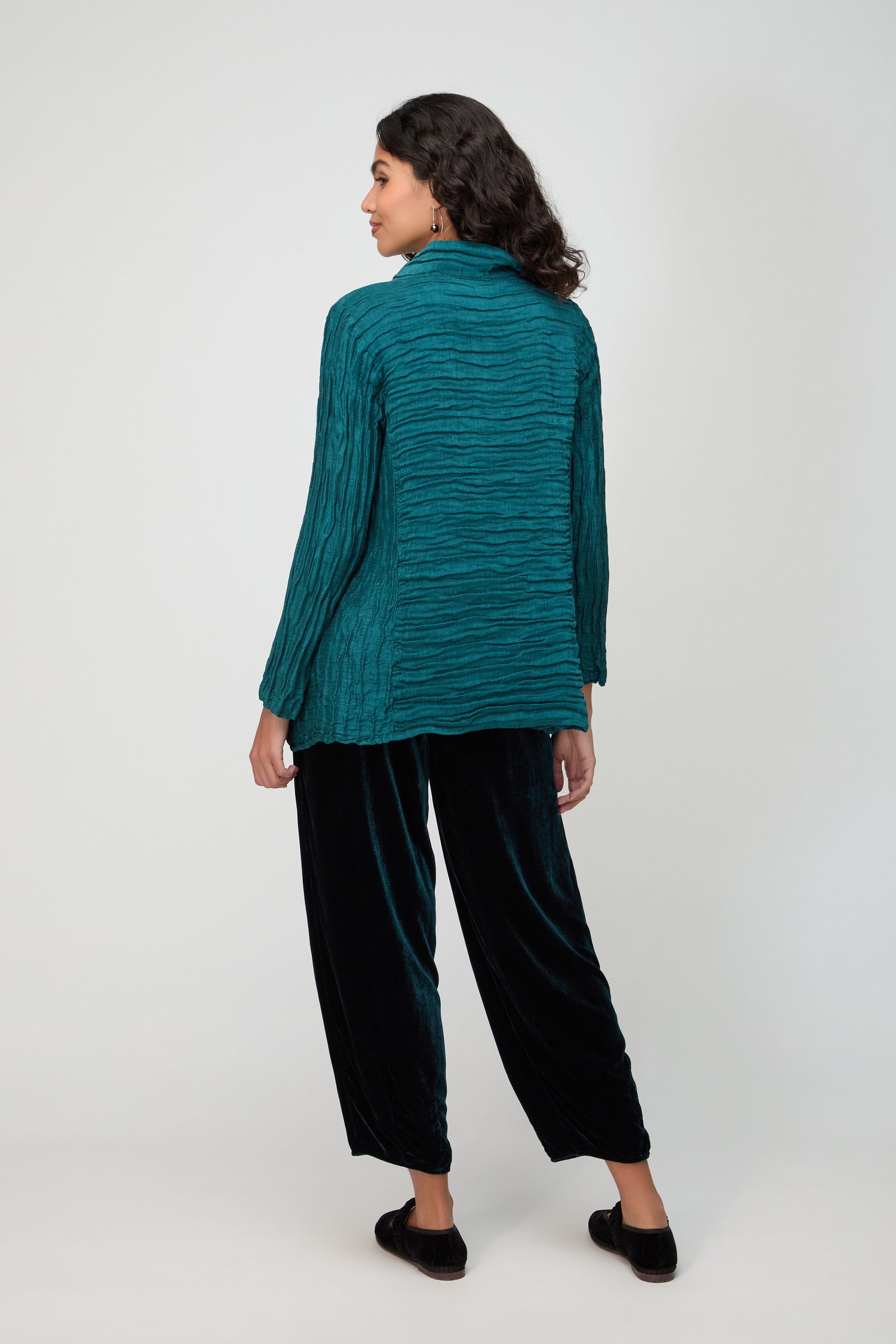 A woman faces away, wearing a teal Panel Front Silk Linen Jacket with black loose-fitting pants and black shoes, set against a plain light background.