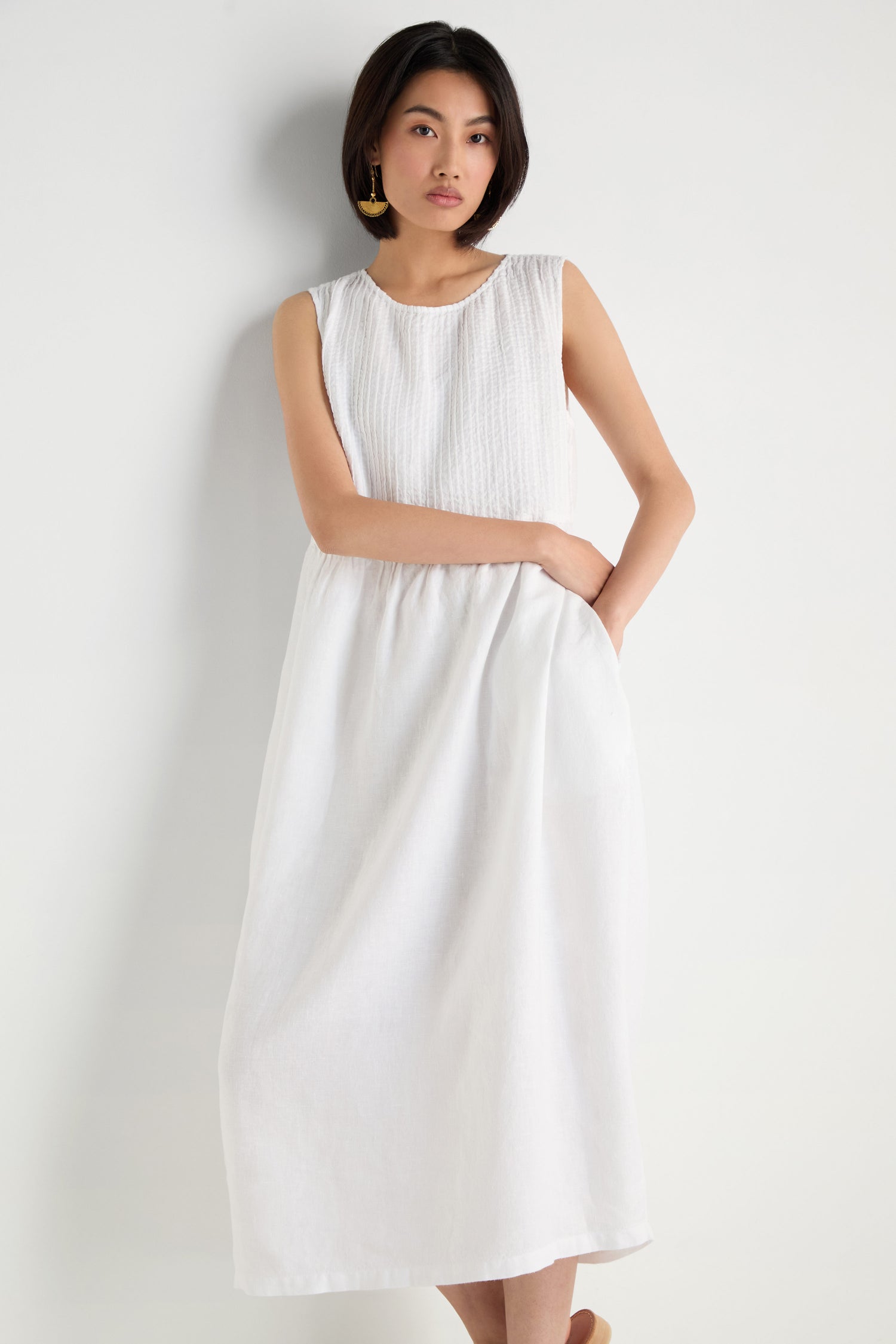 A woman stands against a white background wearing the Pleat Front Linen Bubble Dress, an ankle-length, sleeveless linen dress with textured fabric and gold earrings, with one hand in her pocket.