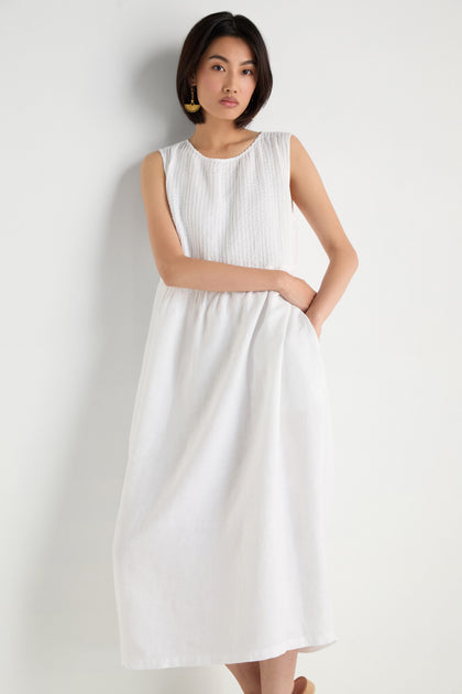 A woman stands against a white background wearing the Pleat Front Linen Bubble Dress, an ankle-length, sleeveless linen dress with textured fabric and gold earrings, with one hand in her pocket.