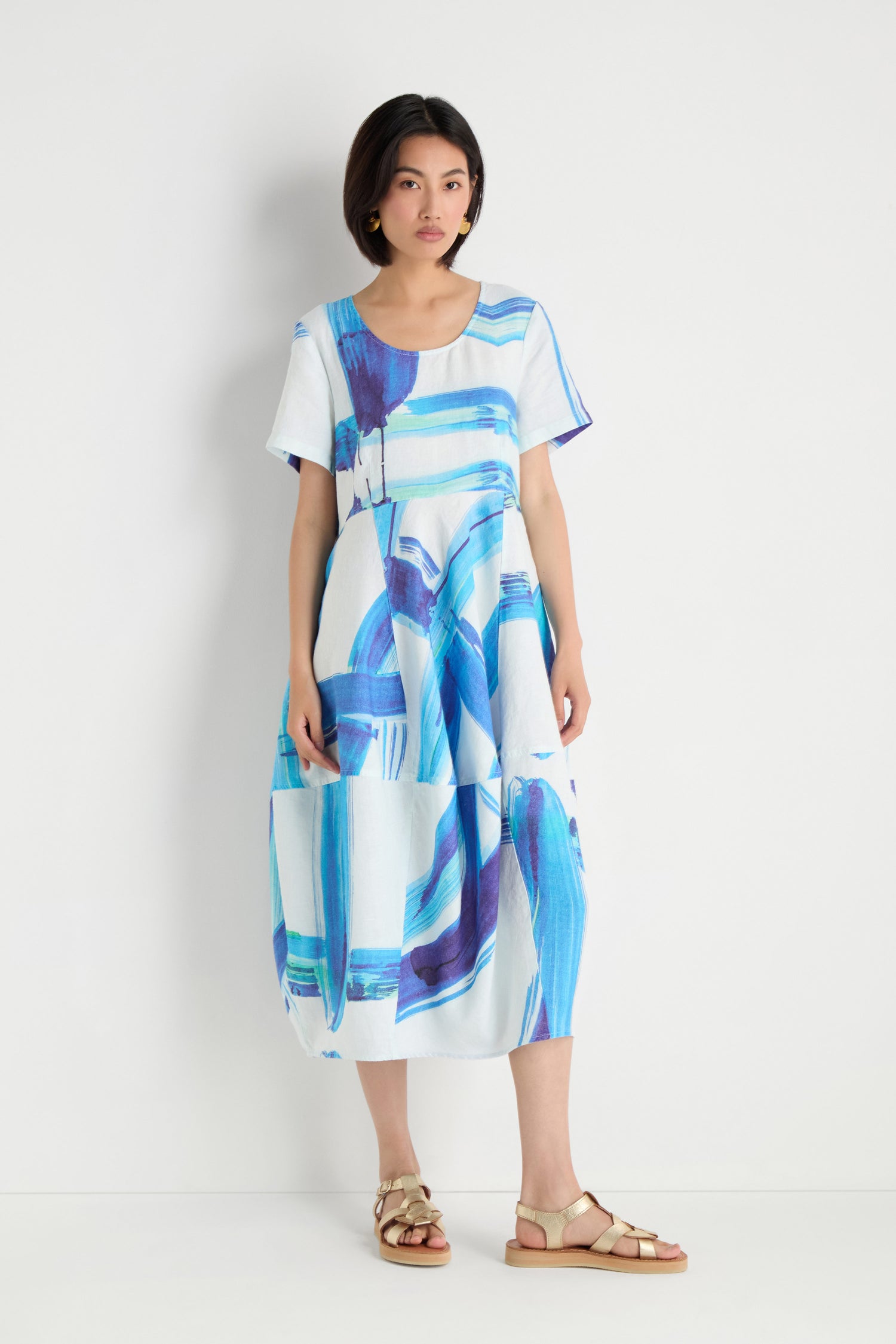 A woman stands against a white wall wearing the Brushstroke Linen Bubble Dress, a loose-fitting linen piece with blue and purple brushstroke print, paired with gold sandals.