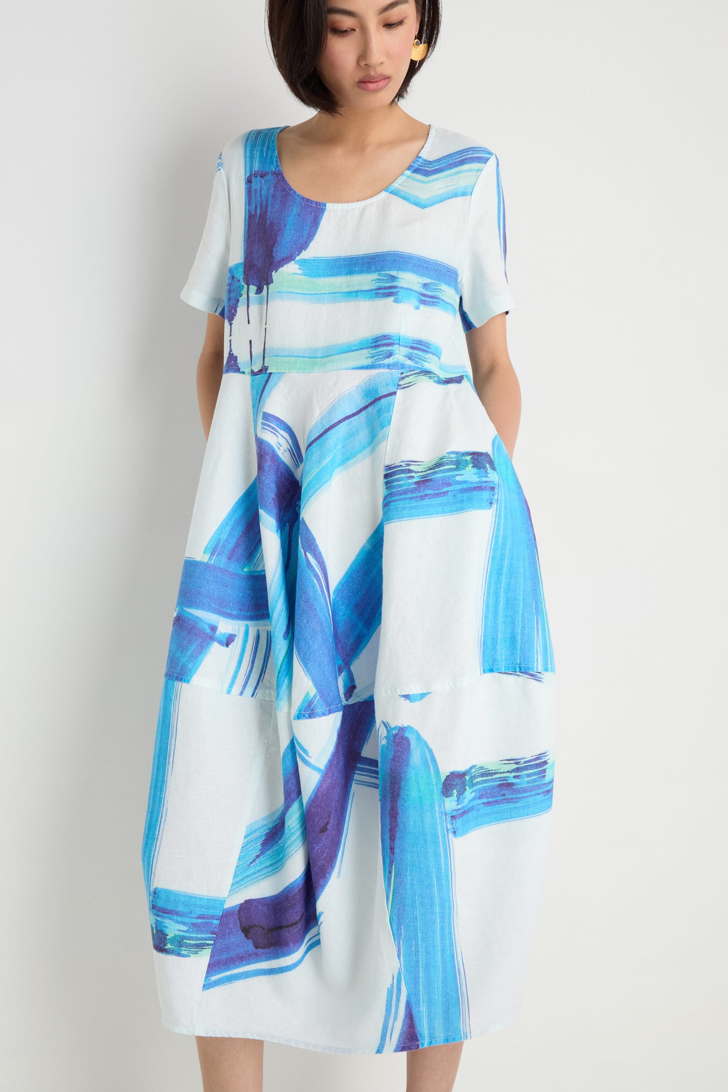 A woman wears the Brushstroke Linen Bubble Dress, featuring blue and purple abstract patterns on white linen, standing against a plain white background.