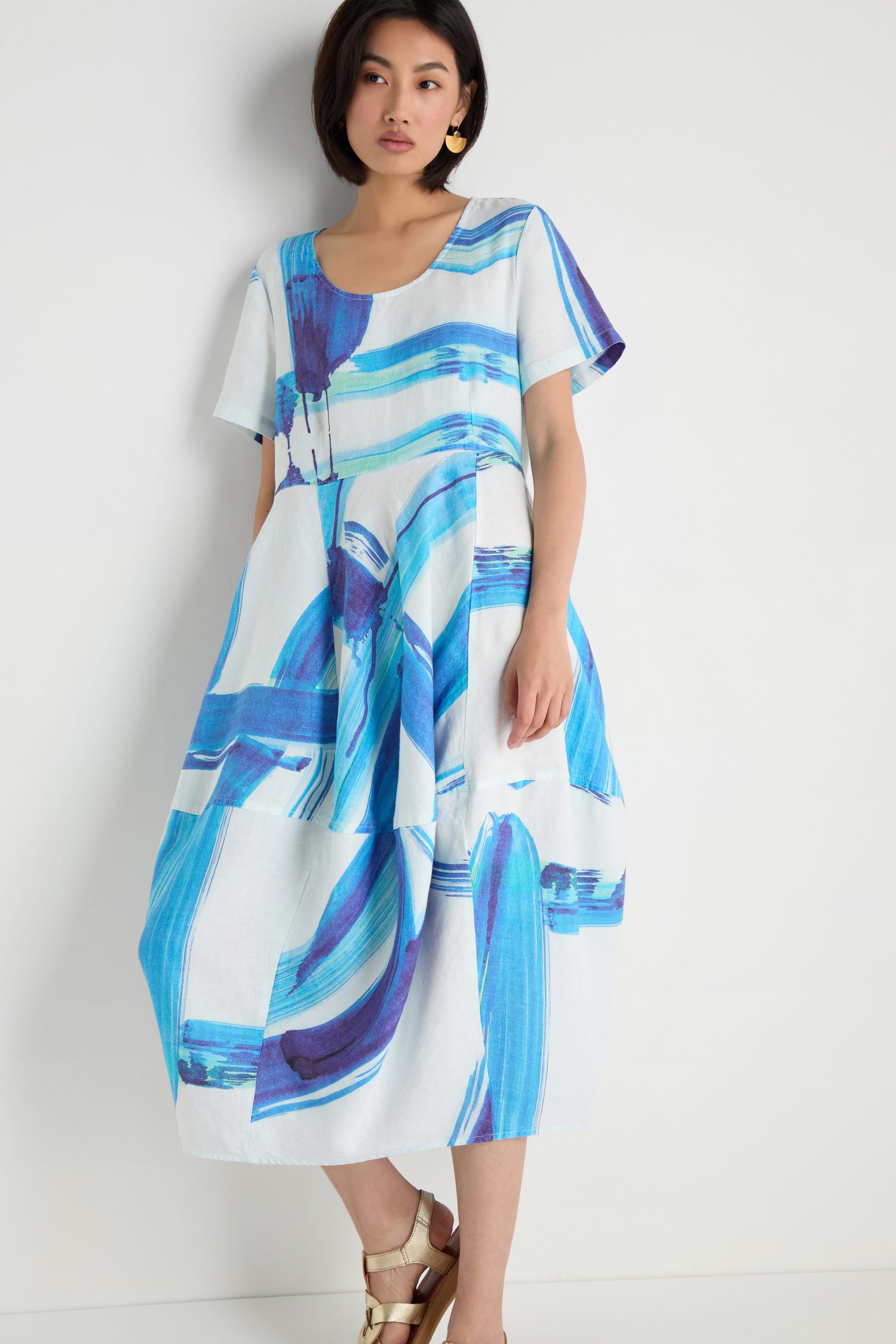 A woman stands against a white wall in the Brushstroke Linen Bubble Dress—white linen with loose short sleeves and blue and purple brushstroke print—paired with gold sandals.