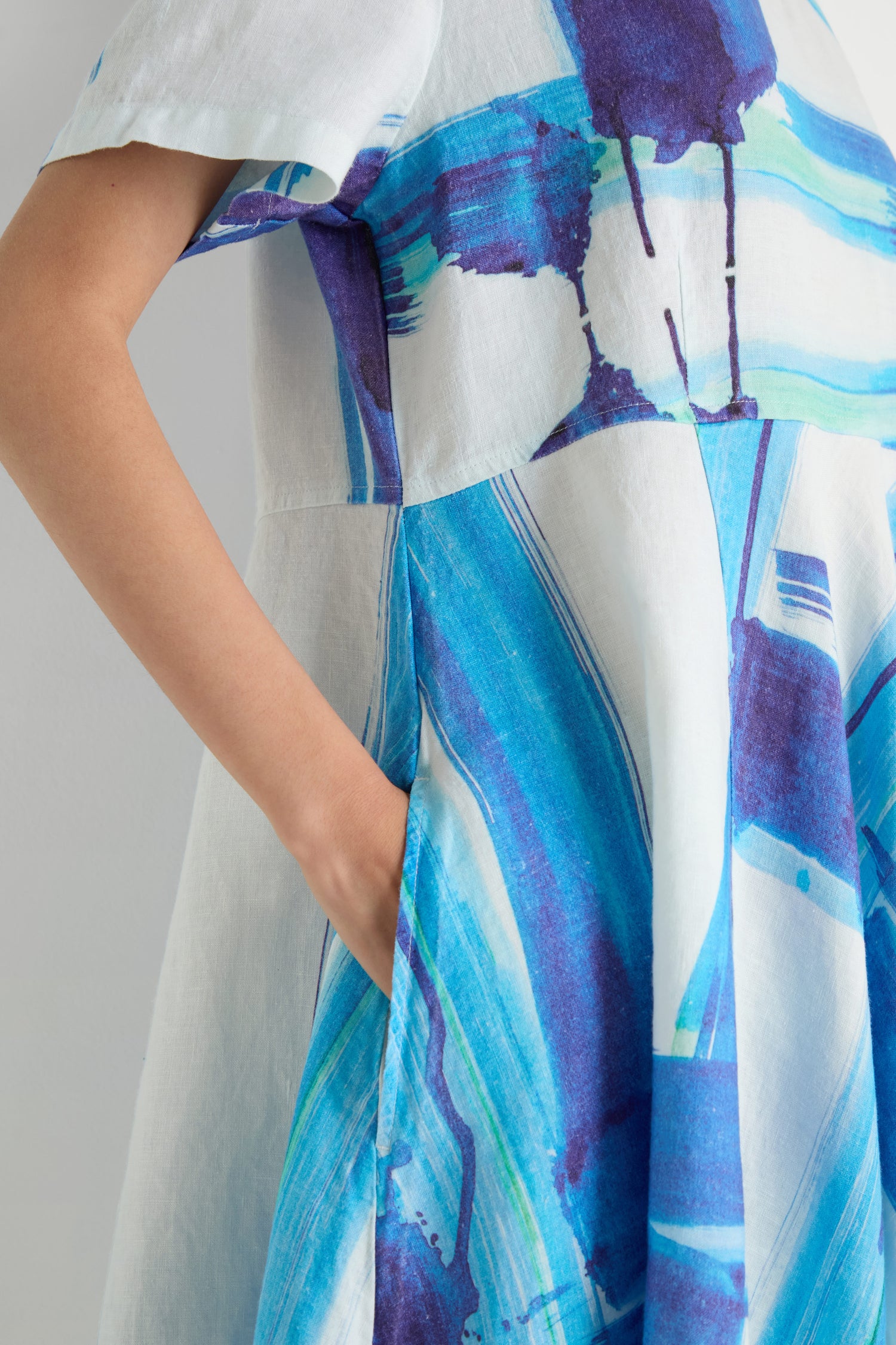 Someone stands with one hand in their pocket, wearing the Brushstroke Linen Bubble Dress featuring a blue and purple print.