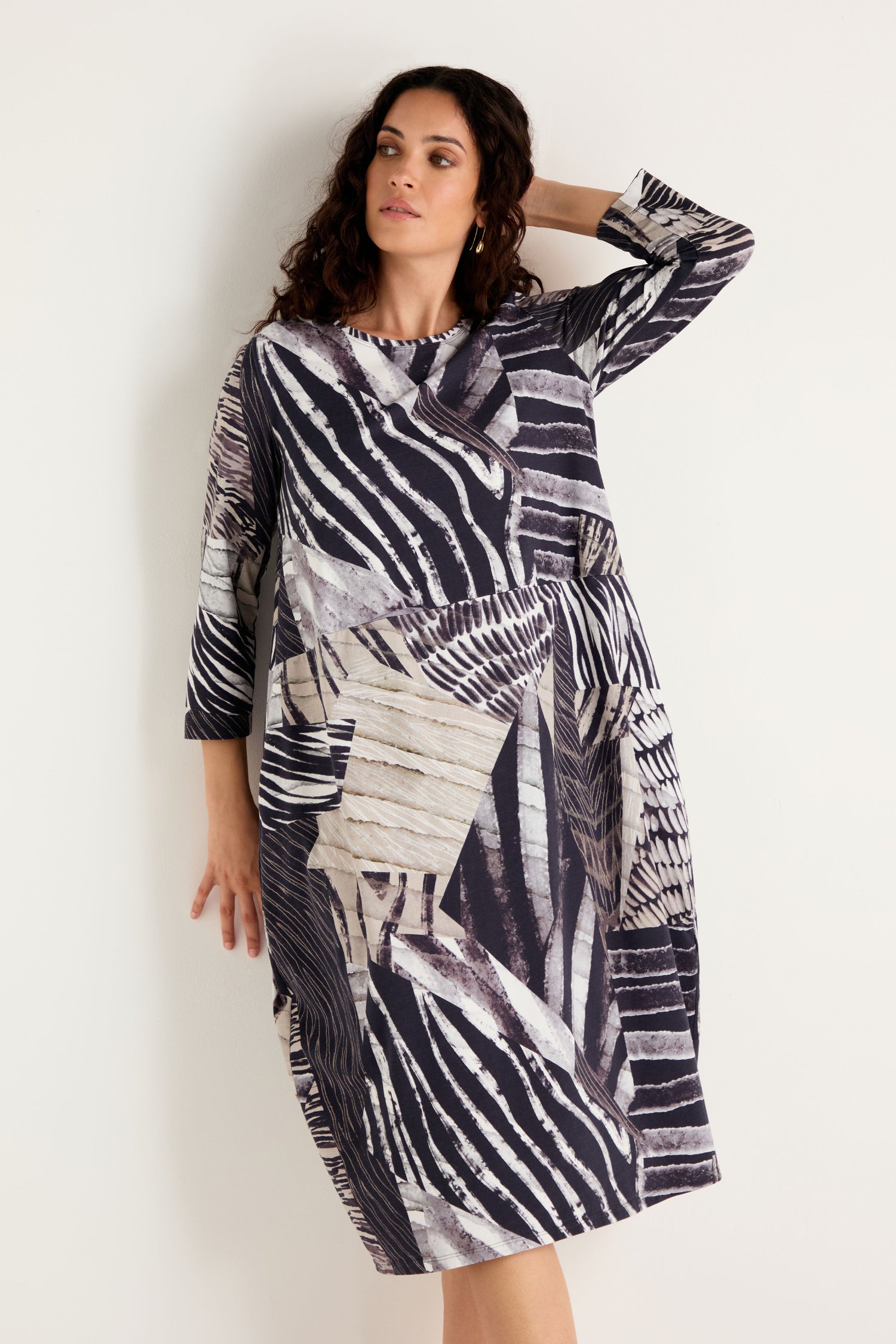 A woman wearing the Monochrome Patchwork Jersey Dress—a loose-fitting, long-sleeve dress featuring an abstract geometric and striped pattern in black, white, and beige—stands against a plain light-colored wall.
