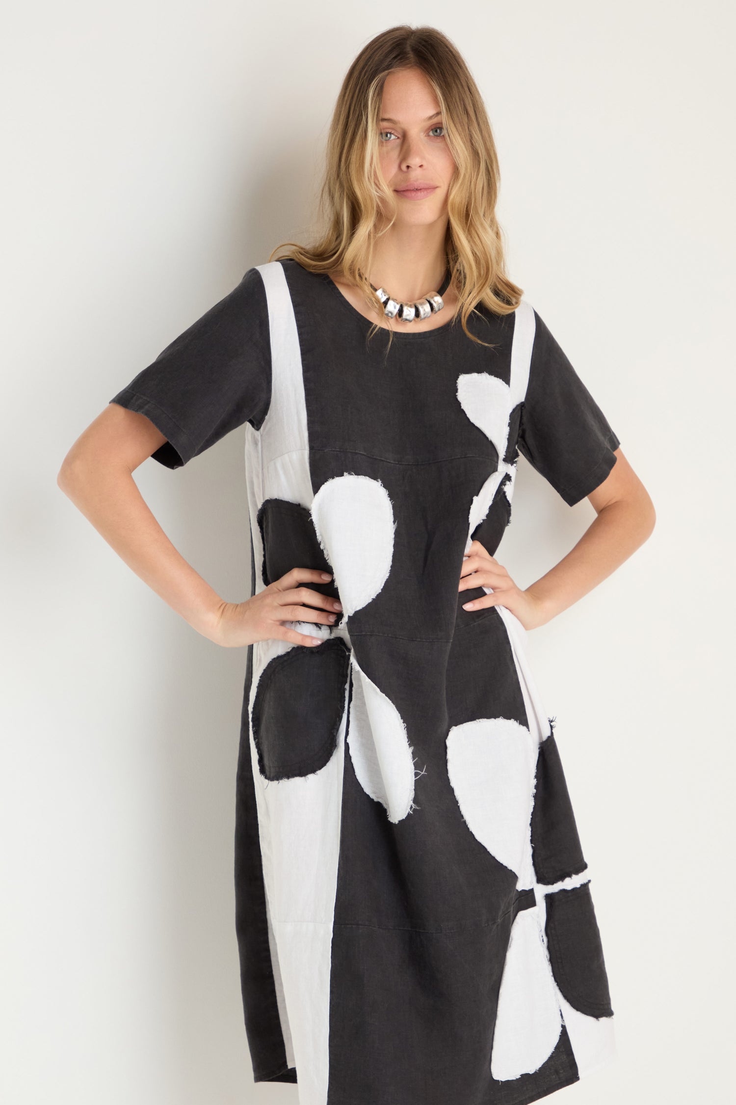 A woman models the Quant Monochrome Linen Dress, a loose-fitting piece featuring large abstract shapes, standing with hands on hips against a plain light background.