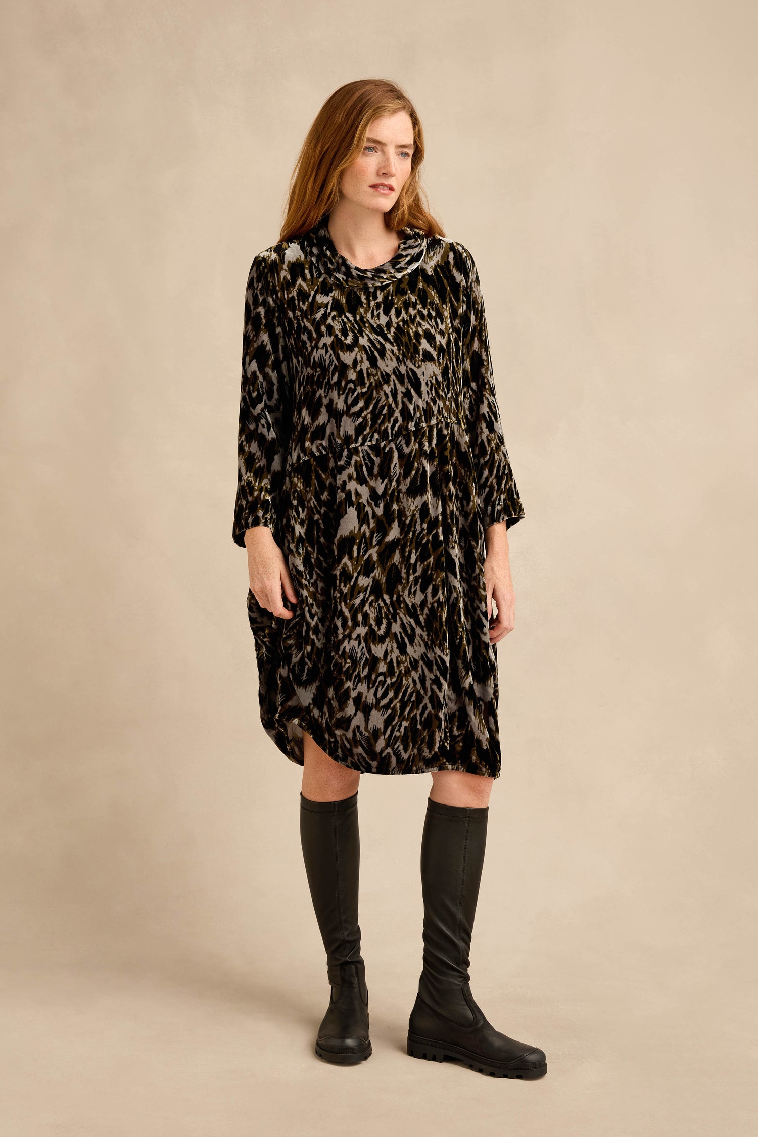 A woman stands against a plain background, wearing the Animal Print Cowl Neck Velvet Dress—a loose, knee-length velvet dress featuring an eye-catching animal print, long sleeves, and a stylish cowl neck—paired with black knee-high boots.