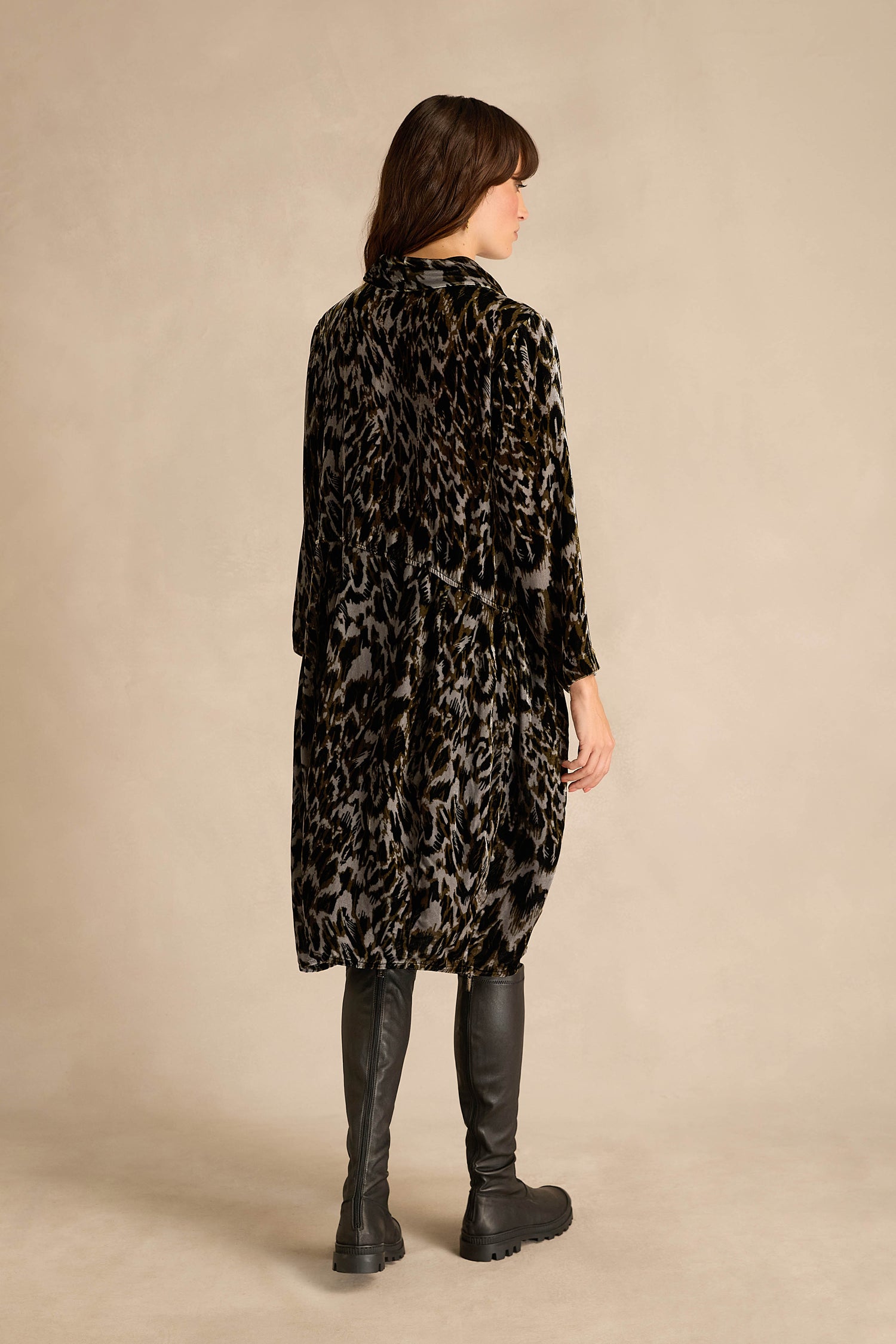 A person with long auburn hair stands facing away, wearing the Animal Print Cowl Neck Velvet Dress—loose-fitting and knee-length—paired with black knee-high boots against a plain beige background.