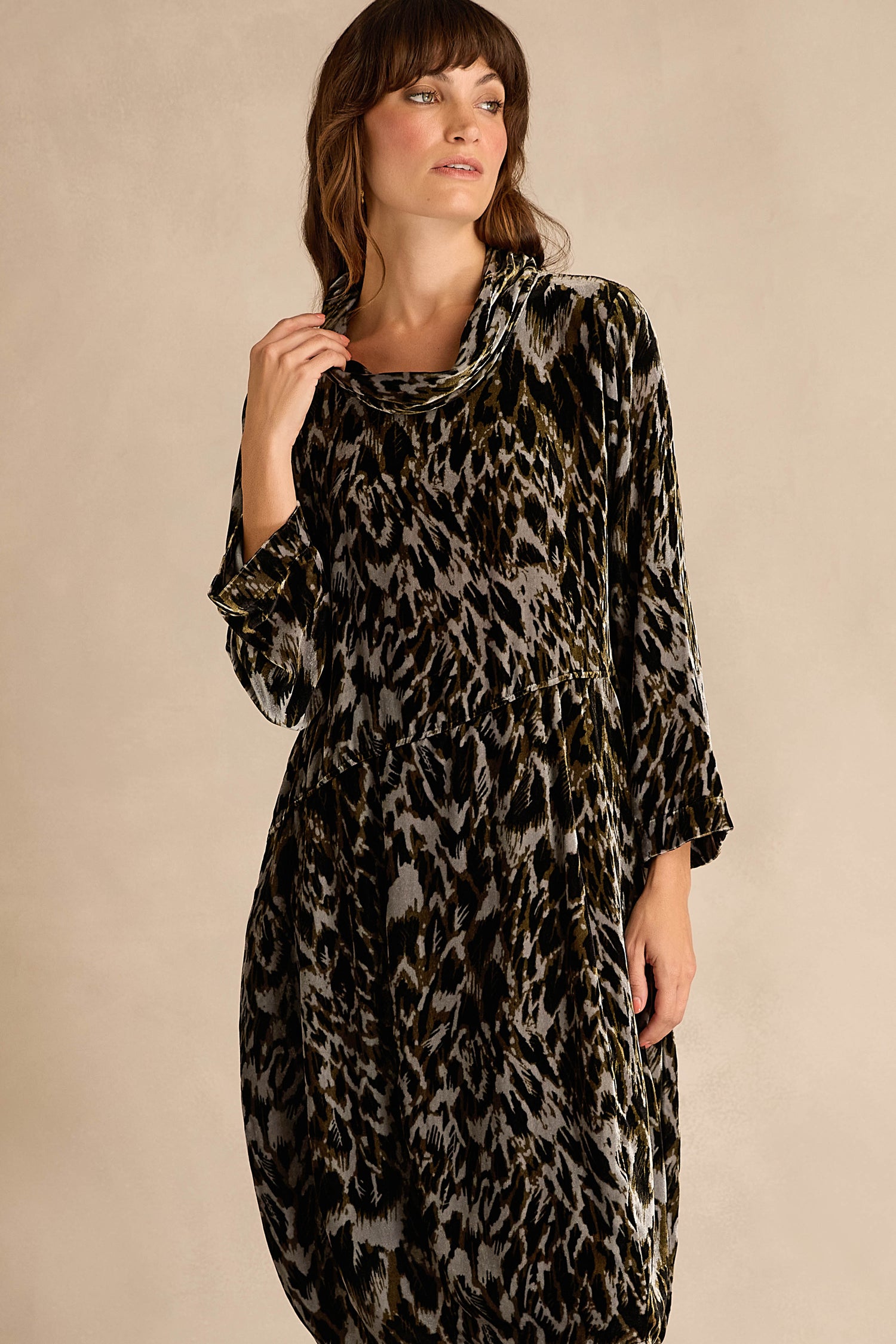 A woman with long brown hair sits on a wooden chair against a beige background, wearing the Animal Print Cowl Neck Velvet Dress.