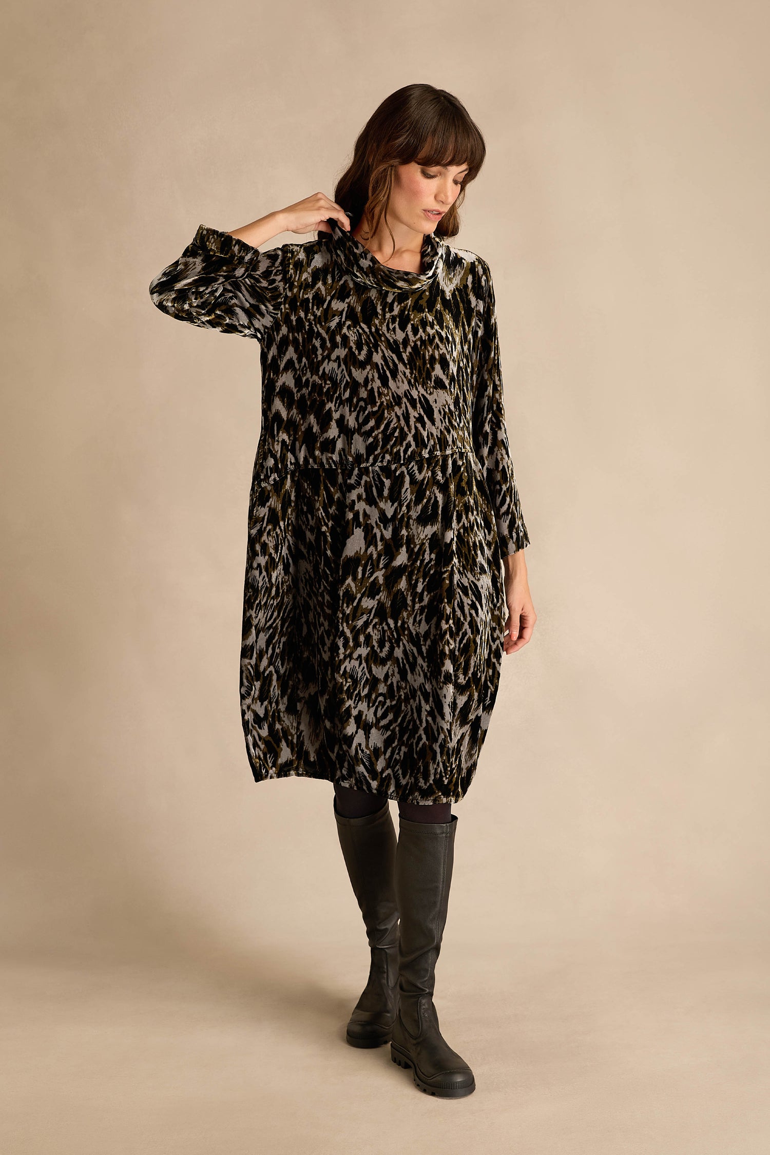 A woman stands against a beige background with one hand in her pocket, wearing the Animal Print Cowl Neck Velvet Dress—a long-sleeved, knee-length dress featuring a chic cowl neckline and stylish animal print.
